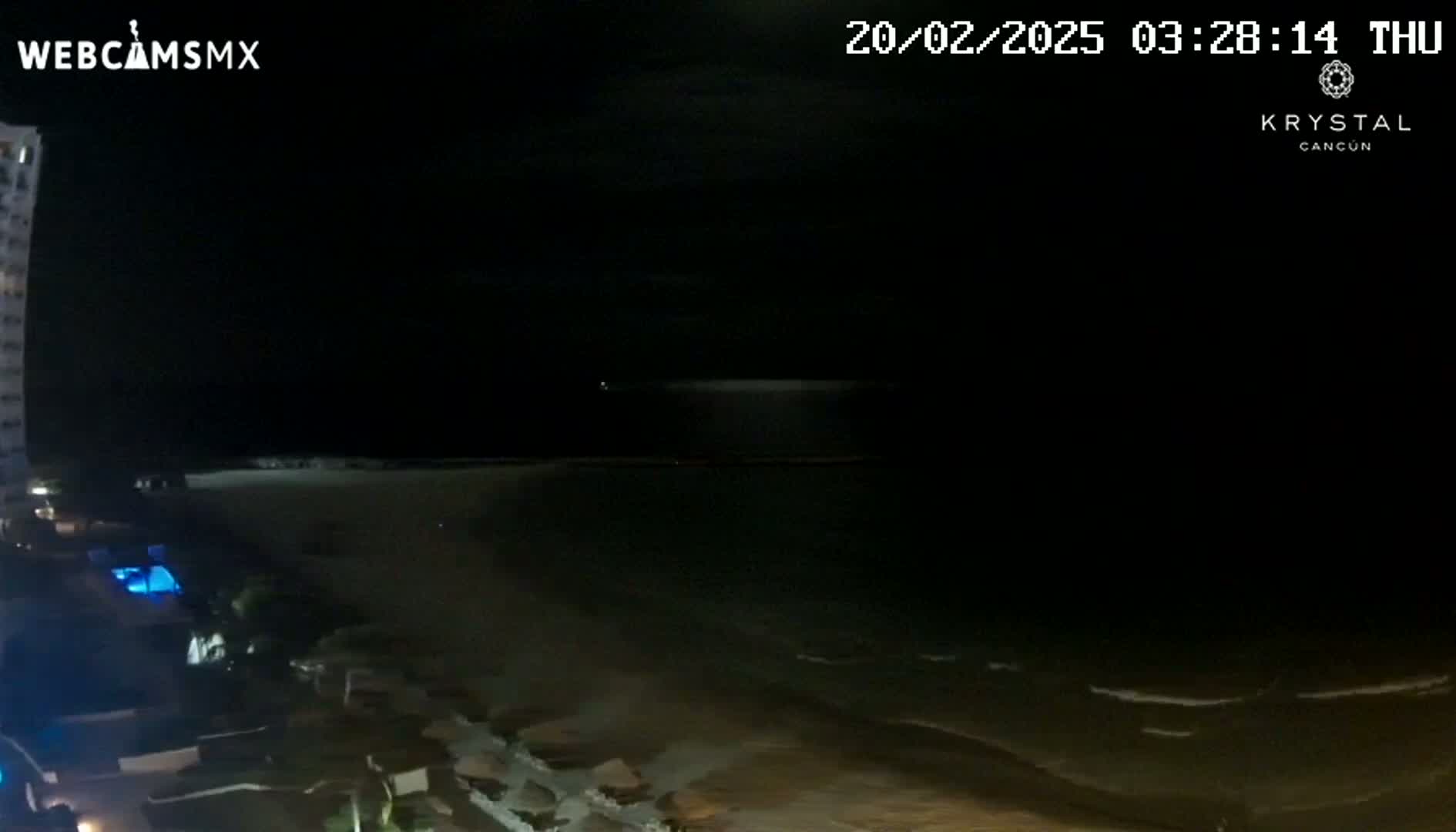 A nighttime view of a beach and ocean under a dark, cloudy sky, with a tall building partially visible in the foreground.