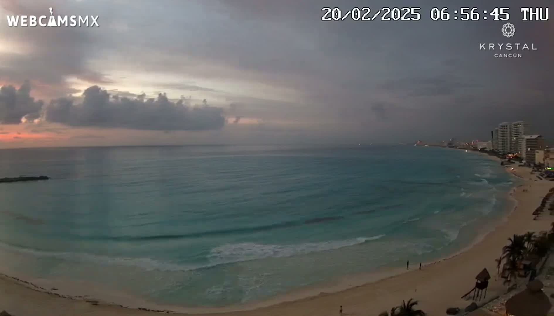 A sandy beach curves along a turquoise ocean under a cloudy, dusky sky at sunrise.