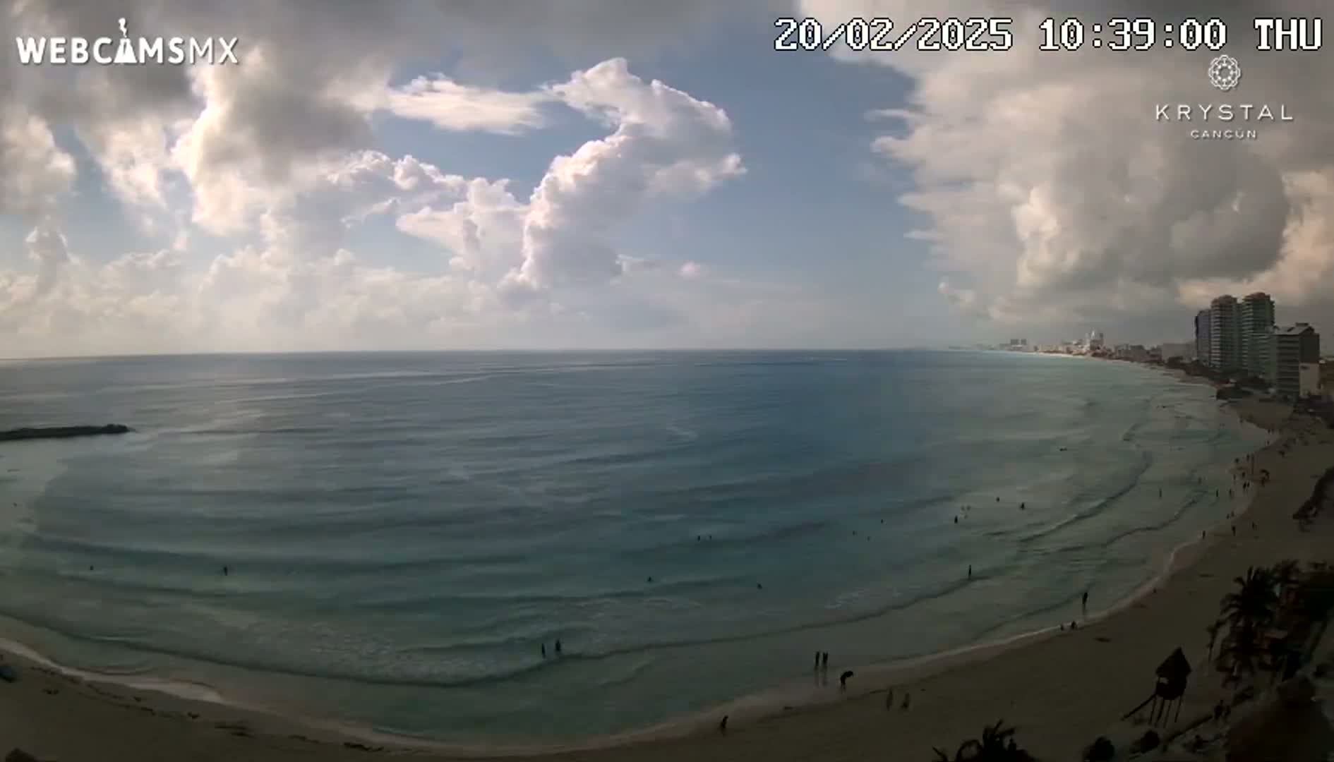 A partly cloudy day shows a long sandy beach with gently rolling waves lapping the shore, and buildings in the distance.