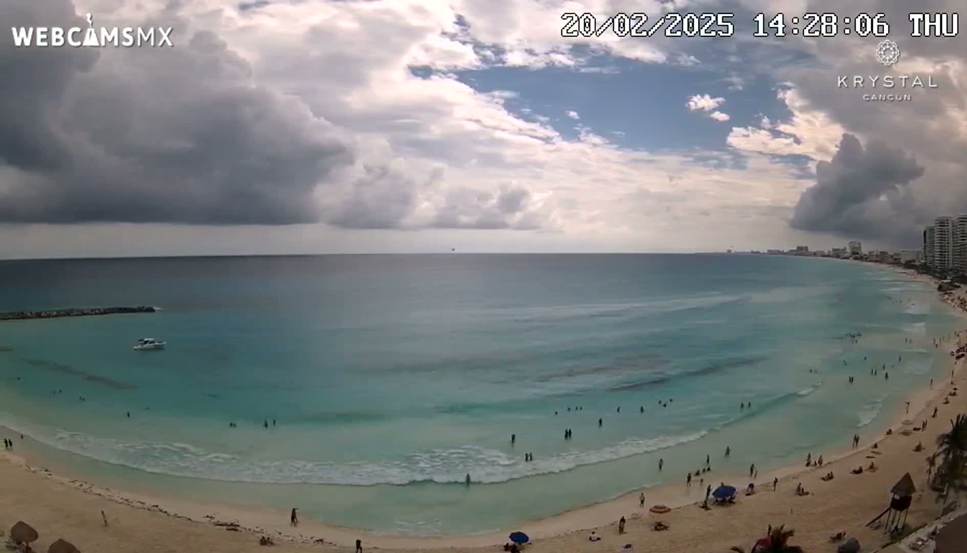 A partly cloudy day shows a beach with turquoise water and many people swimming and sunbathing, with buildings visible in the distance.