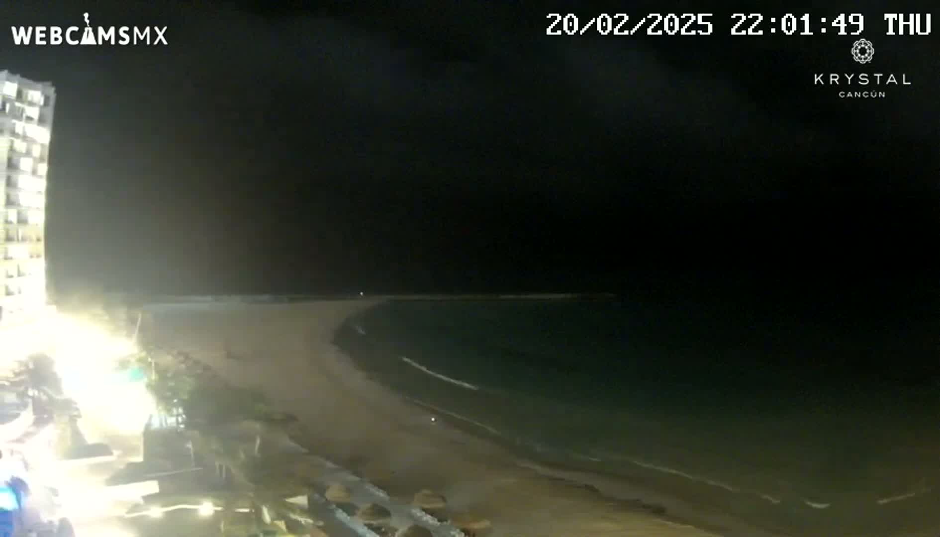 A nighttime view shows a sandy beach curving into a dark ocean under a clear sky, with a tall building illuminated on the left.