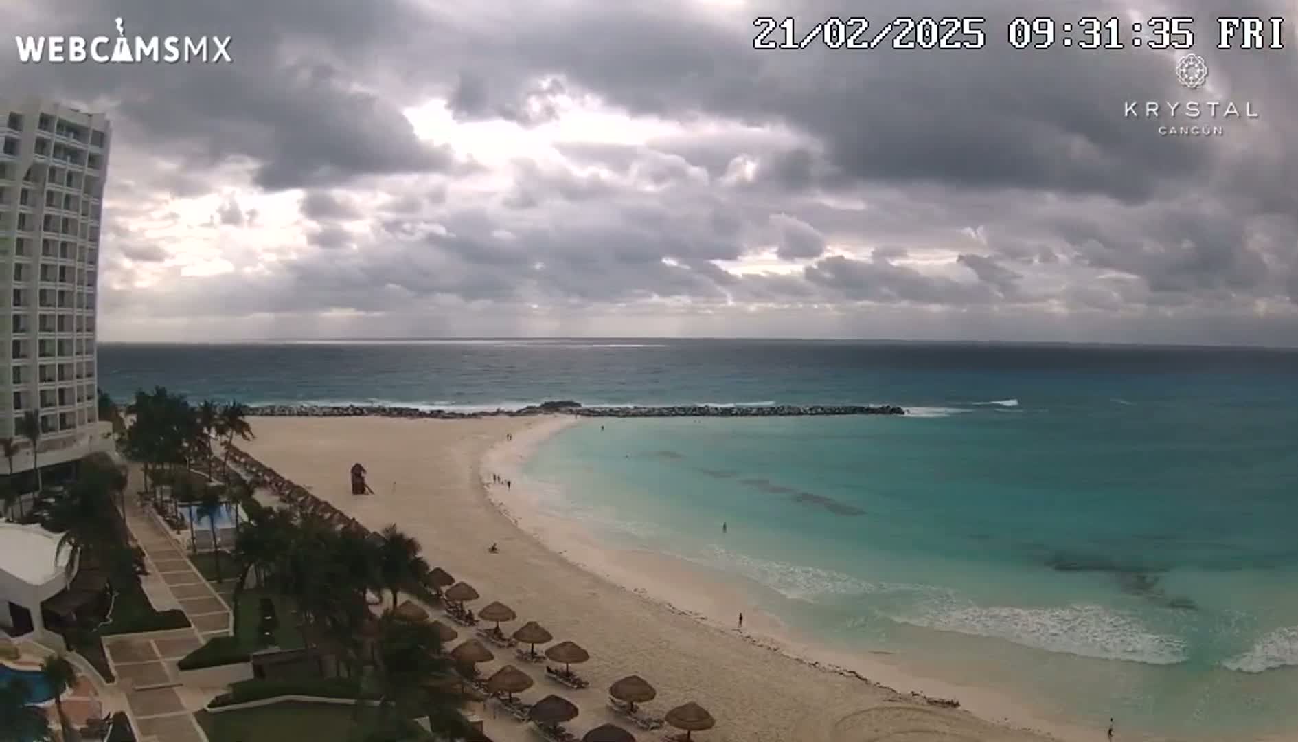 A mostly cloudy day shows a beach with turquoise water, a white sand shoreline, and a tall white hotel overlooking the scene.