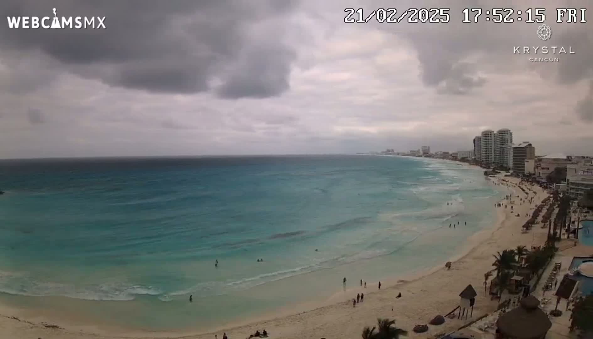 An overcast sky hangs over a beach with turquoise water and a few people scattered along the shore, with high-rise hotels visible in the distance.
