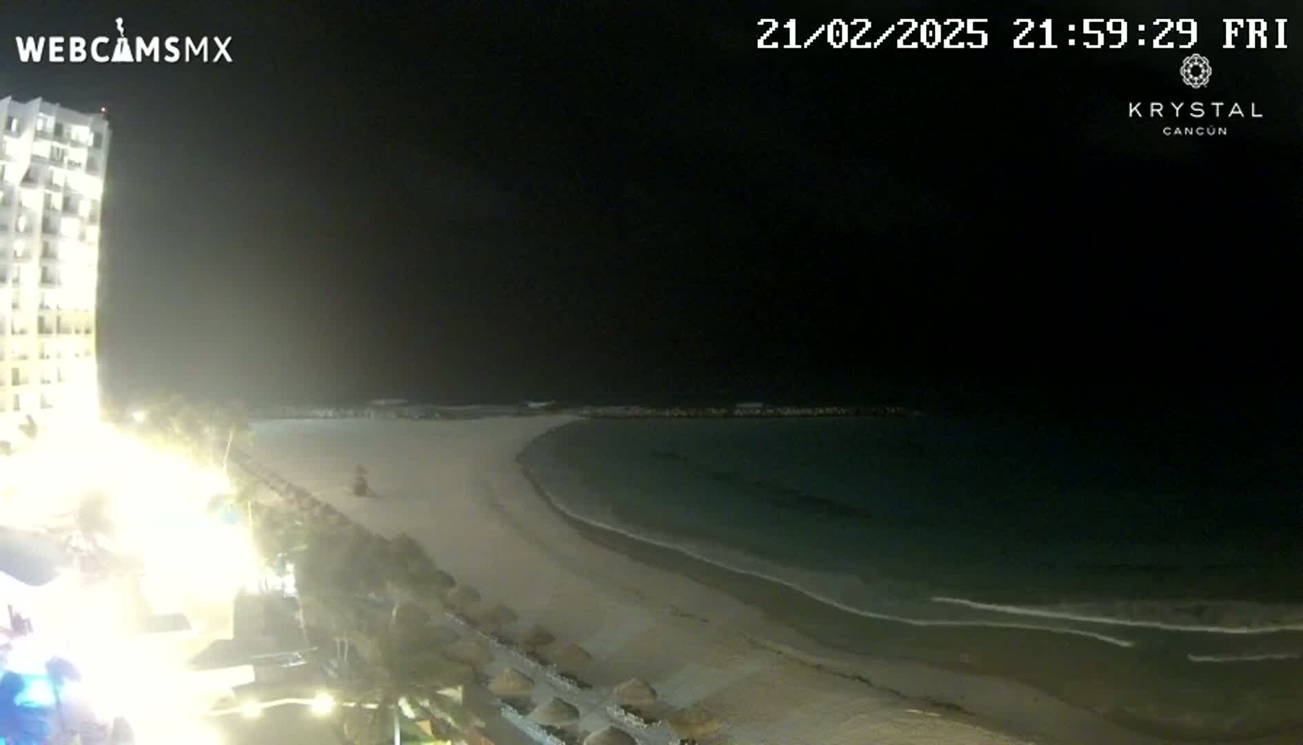 A nighttime view shows a brightly lit beach with calm water and a tall building to the left.