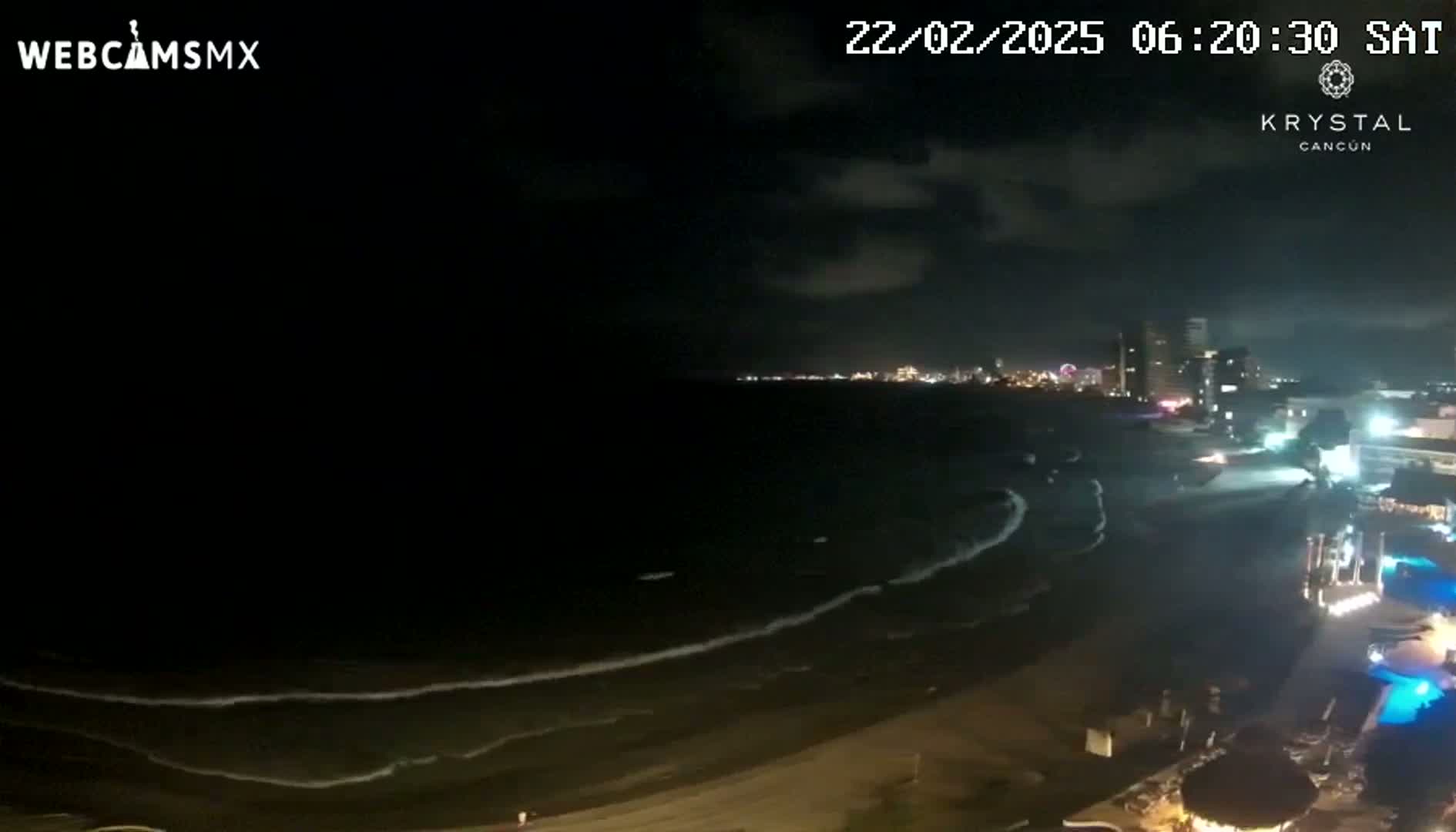 A nighttime beach scene shows gently rolling waves lapping the shore under a partly cloudy sky, with a city skyline visible in the distance.
