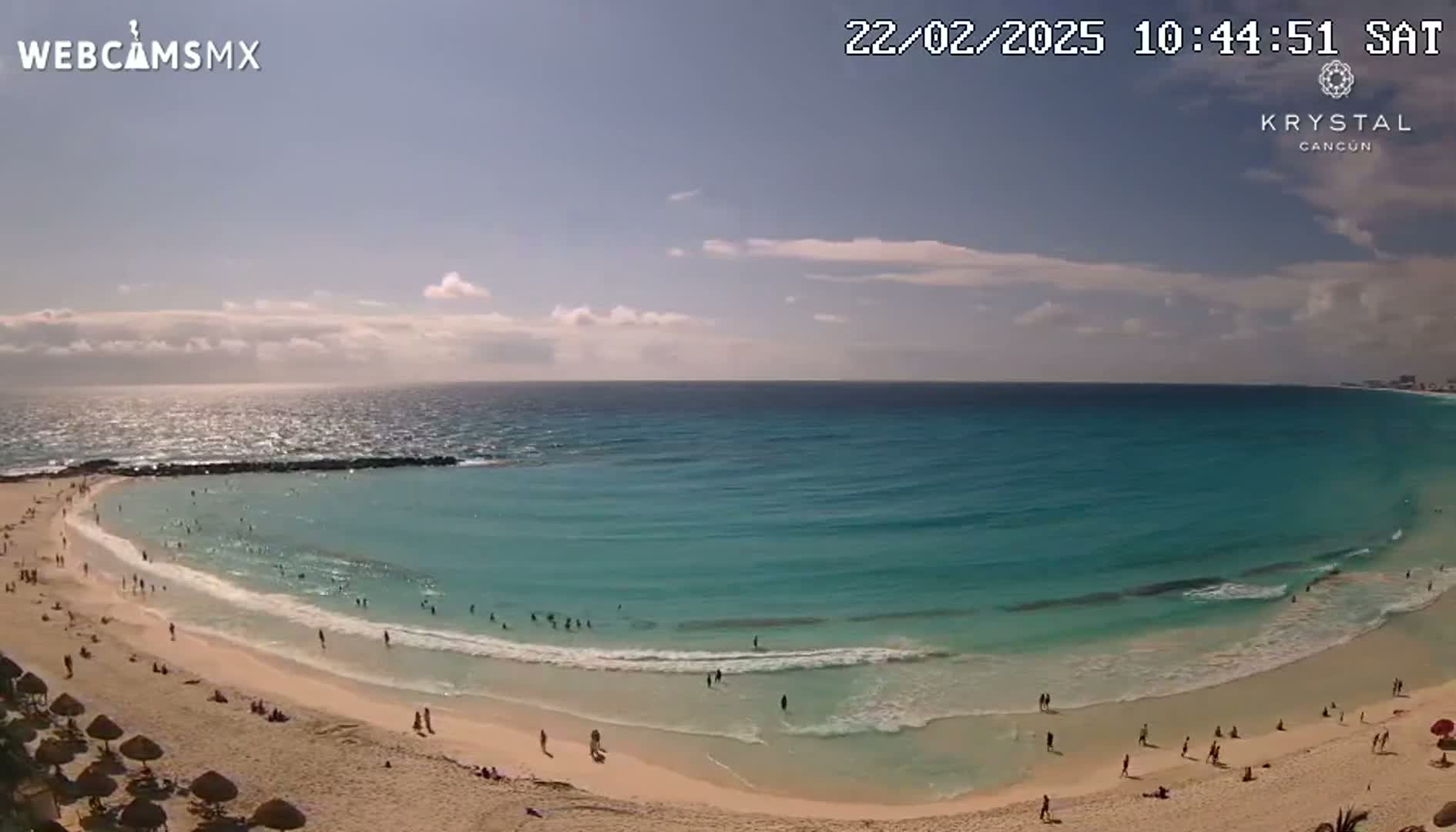 A partly cloudy day shows a sandy beach with turquoise water and a few people enjoying the sun.