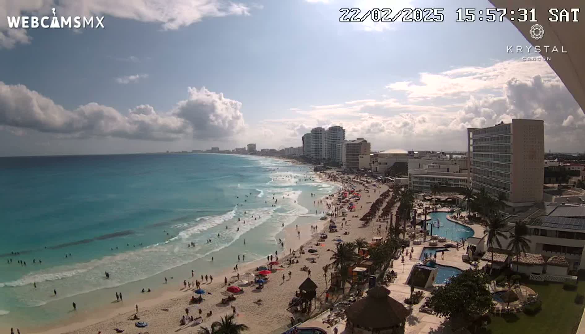 A partly cloudy day at a beach resort shows a crowded beach with turquoise water and waves, along with multiple high-rise hotels and palm trees.