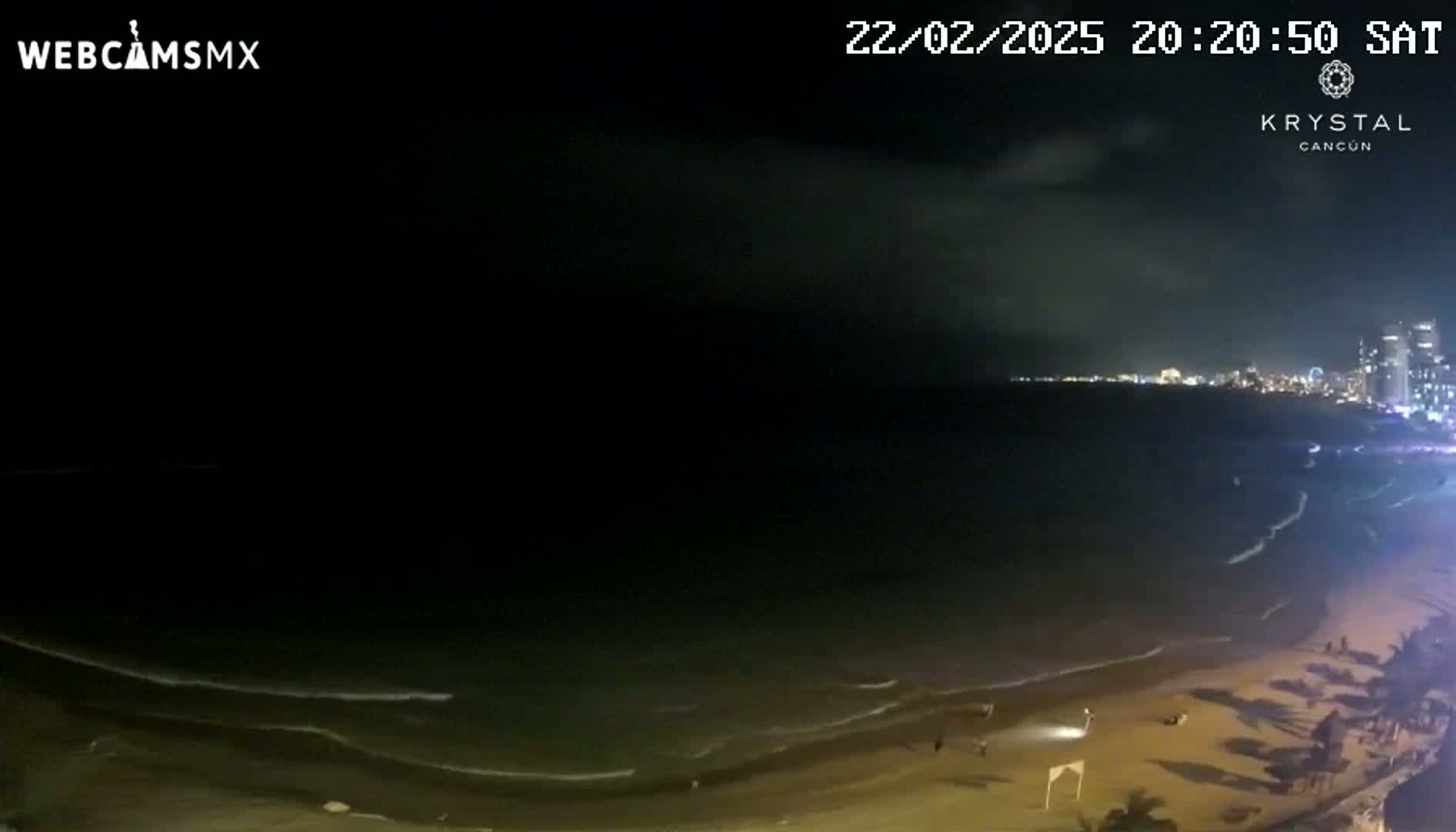 A nighttime beach scene shows dark water with small waves lapping onto a sandy shore under a cloudy sky.