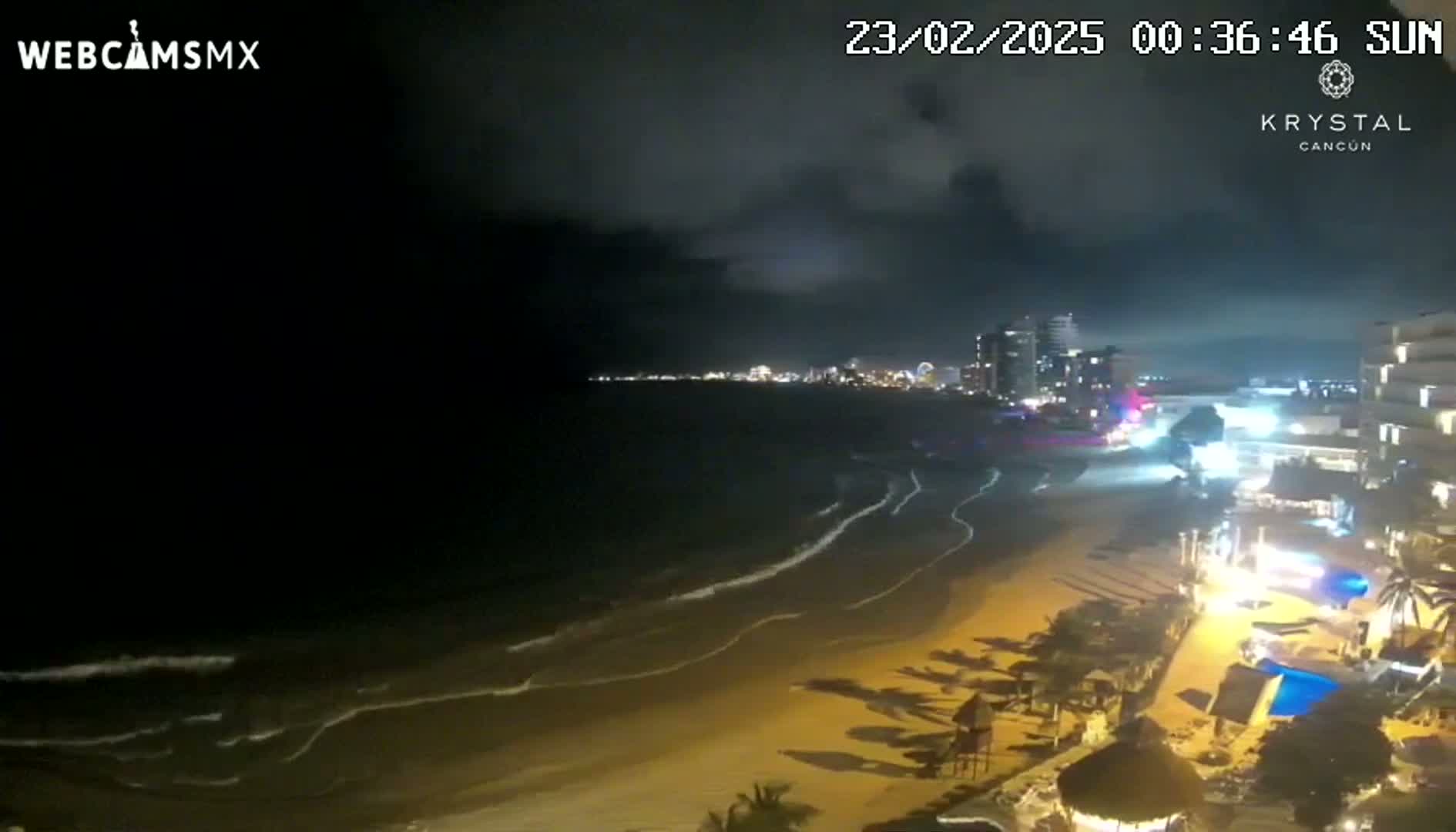 A nighttime beach scene shows a dark, cloudy sky over gently lapping waves on a sandy shore, with illuminated buildings and resort features visible along the coastline.