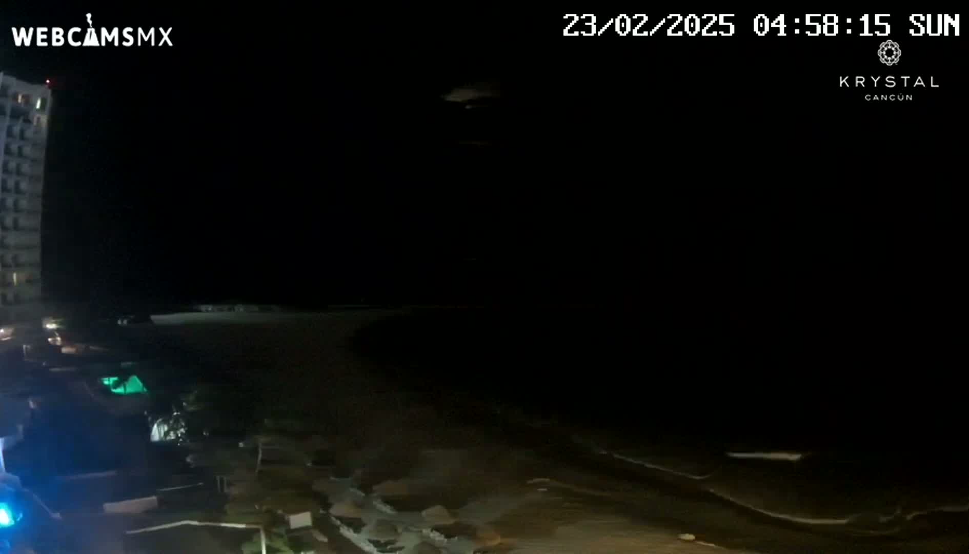 A nighttime view shows a dark beach with small waves lapping the shore near a tall building under a clear, dark sky.