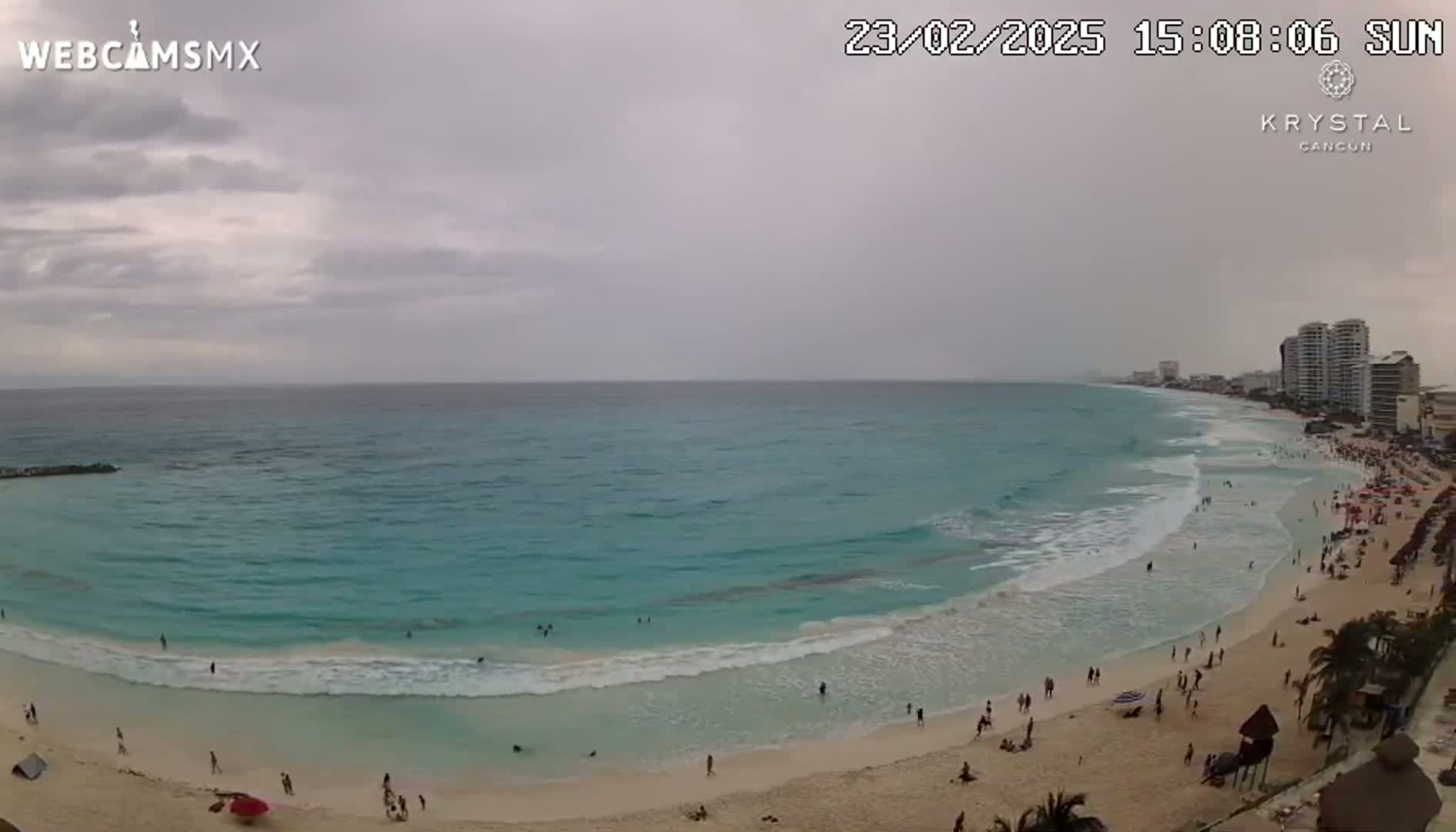A beach with turquoise water and small waves under an overcast sky shows many people on the sand.