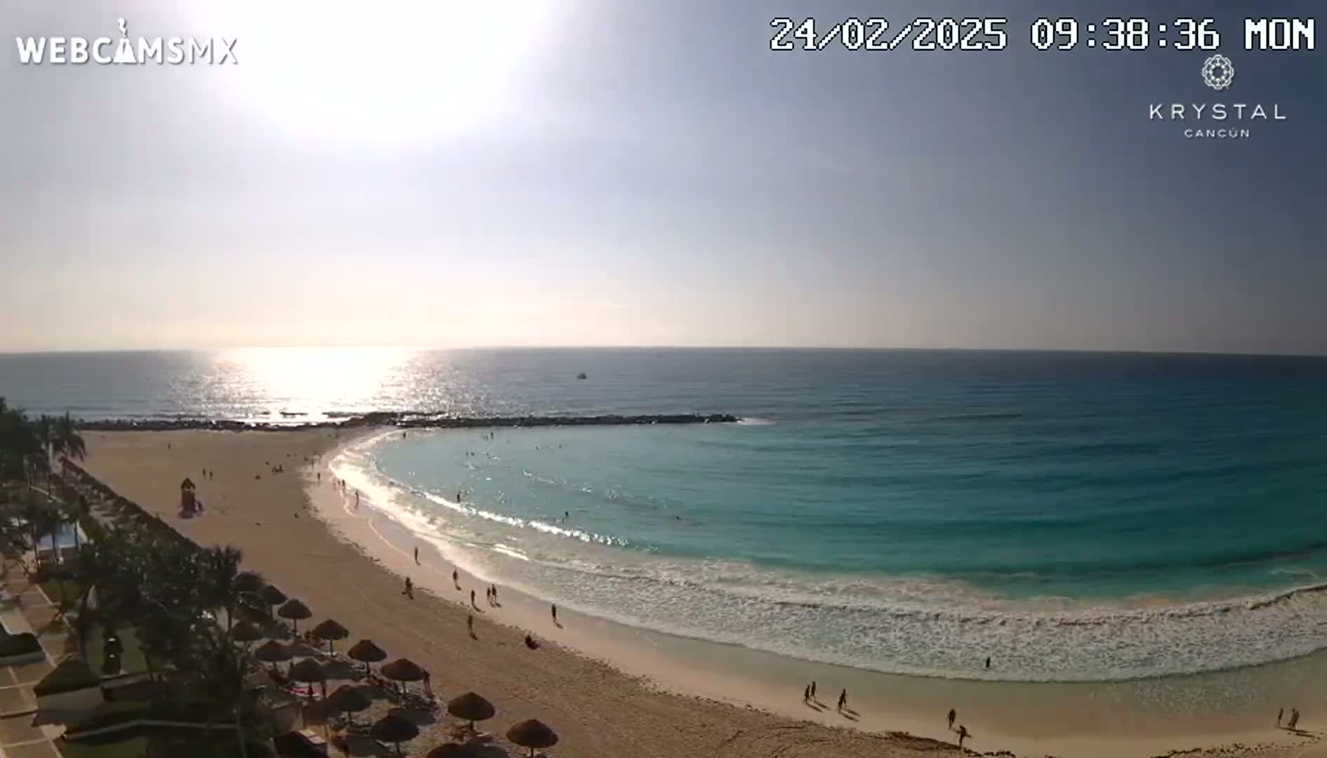 A mostly sunny day at a beach with turquoise water lapping a sandy shore, where a few people are scattered along the sand and in the water.