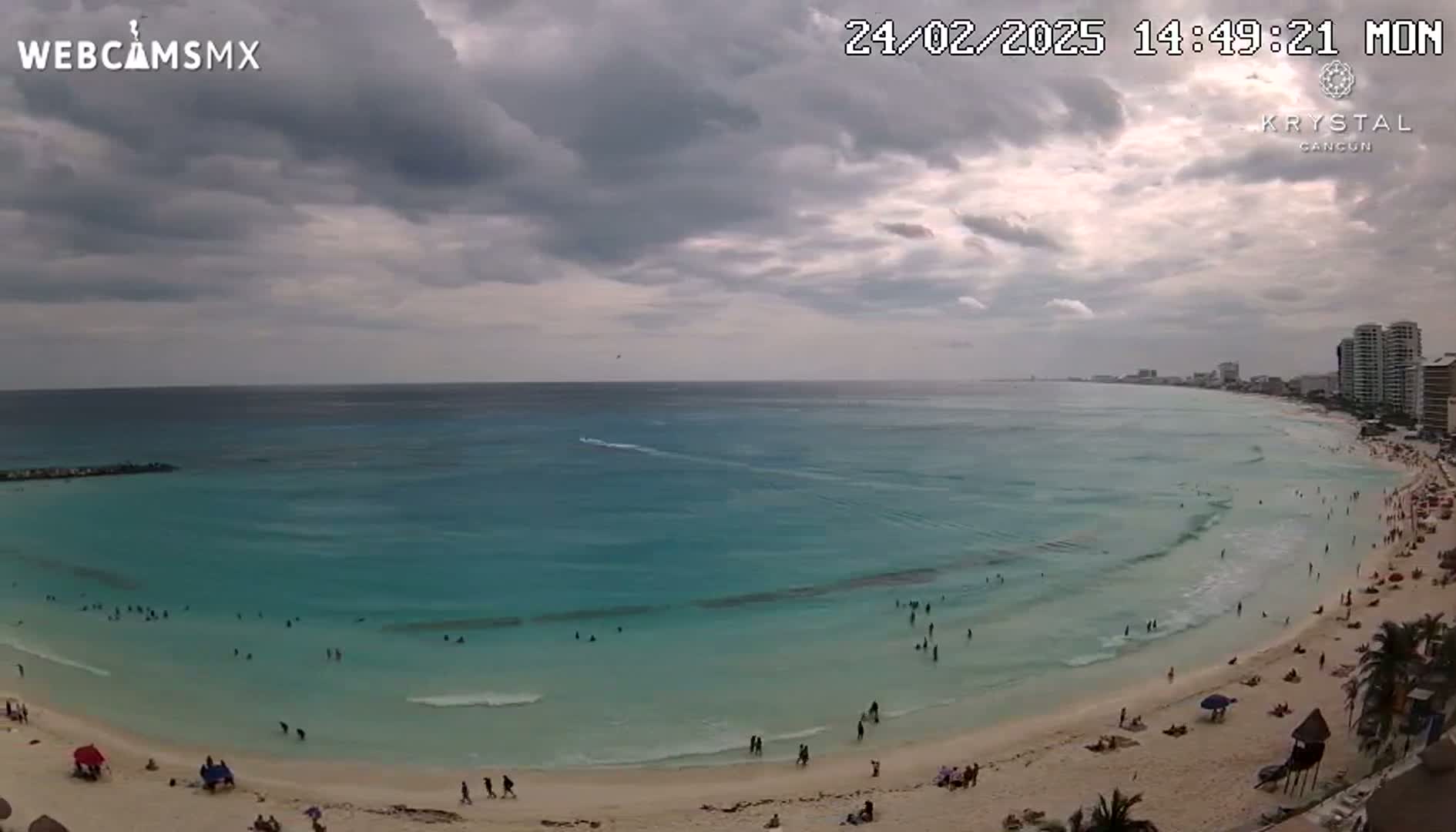 A mostly cloudy sky overlooks a beach with turquoise water and many people swimming and relaxing on the sand.