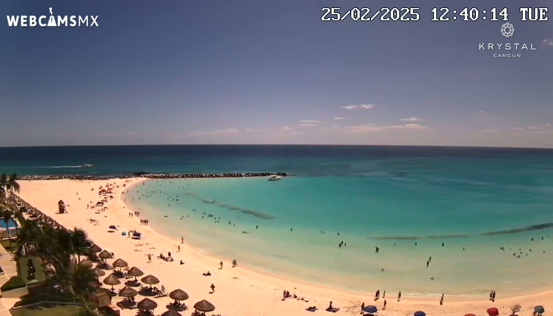 A mostly sunny day shows a beach with turquoise water and many people enjoying the sand and ocean.