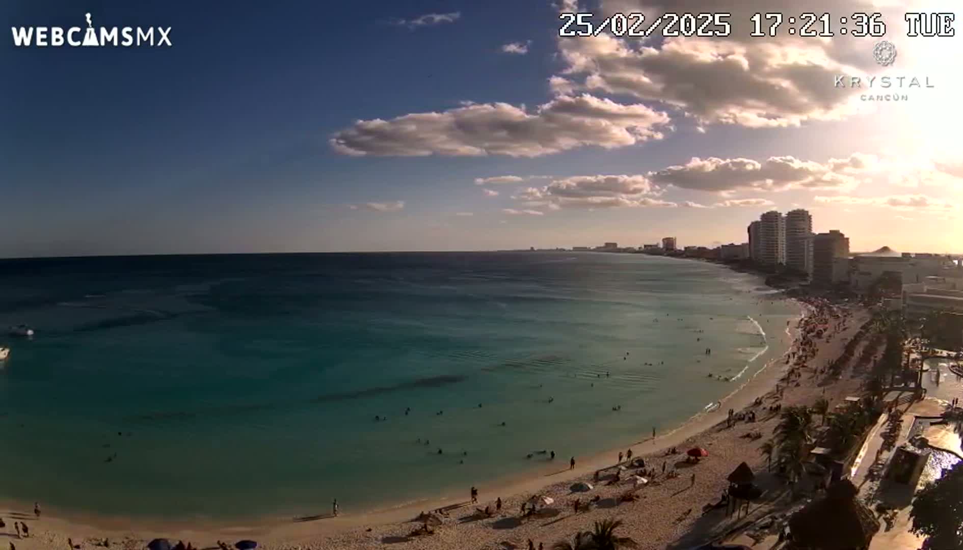 A partly cloudy sunset over a beach with turquoise water and many people enjoying the sand and ocean.