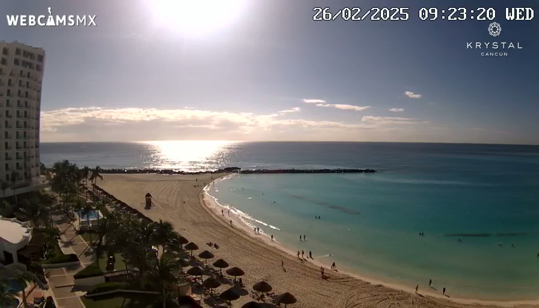 A sunny day shows a sandy beach curving into a calm, turquoise ocean, with a high-rise hotel and palm trees visible to the left.