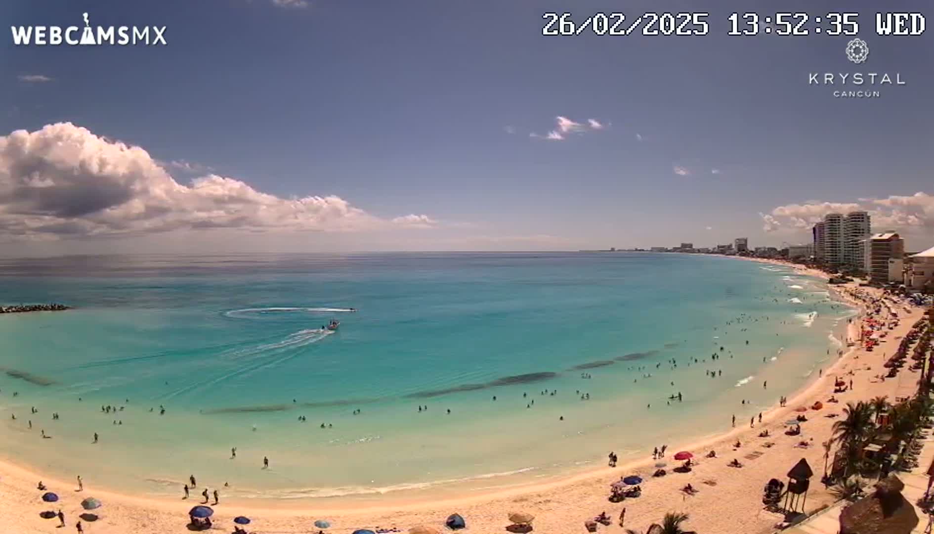 A sunny day on a beach with clear turquoise water,  a sandy shore populated with people and umbrellas, and buildings visible in the distance.