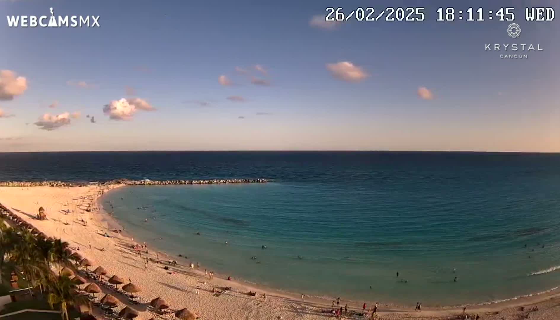 A mostly clear, sunset sky overlooks a sandy beach with calm, turquoise water and a few people swimming and lounging.