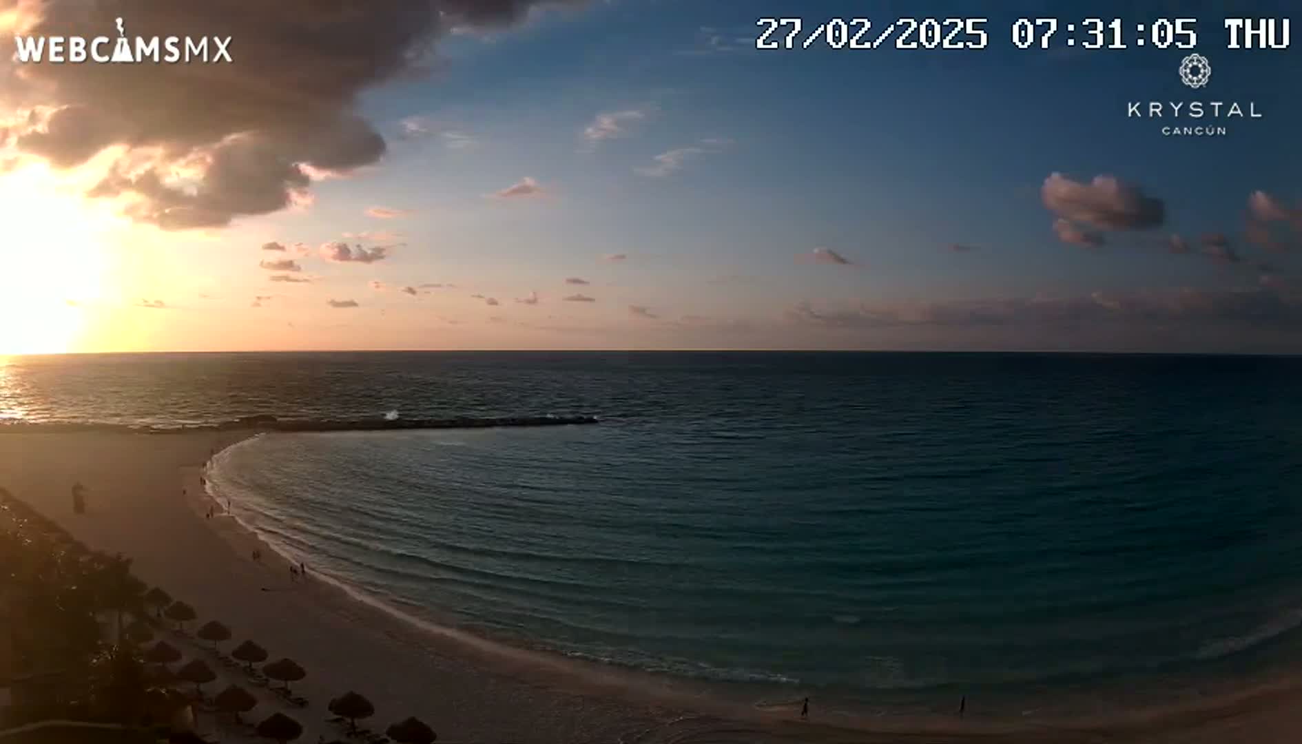 A sunrise over a calm ocean, with a sandy beach and some palm trees visible in the foreground.
