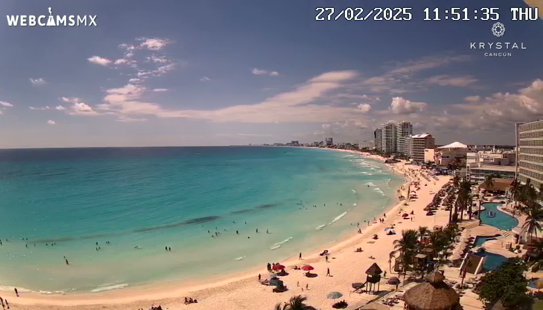 A partly sunny day shows a long sandy beach with turquoise water and many people swimming and relaxing, next to a hotel zone with tall buildings.
