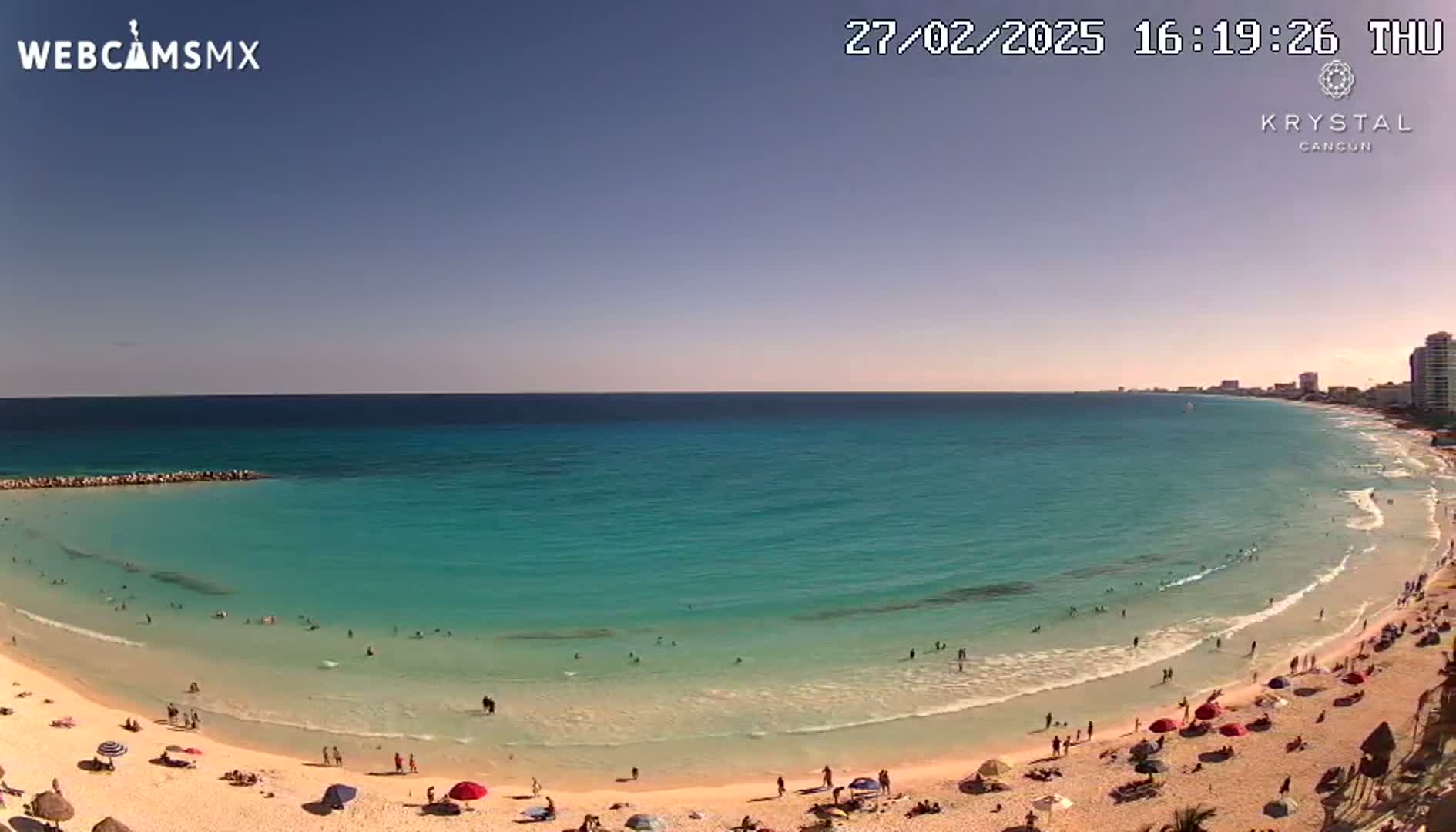 A sunny day at a beach shows a clear turquoise ocean with people swimming and relaxing on a sandy shore,  with buildings visible in the distance.