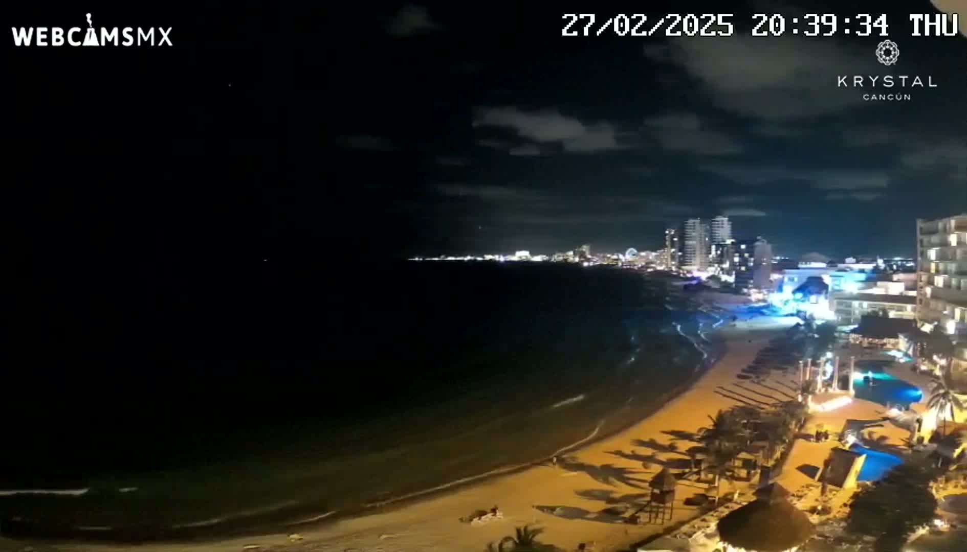 A nighttime beach scene shows a dark ocean with gentle waves lapping a sandy shore, lined with illuminated buildings and palm trees under a partly cloudy sky.
