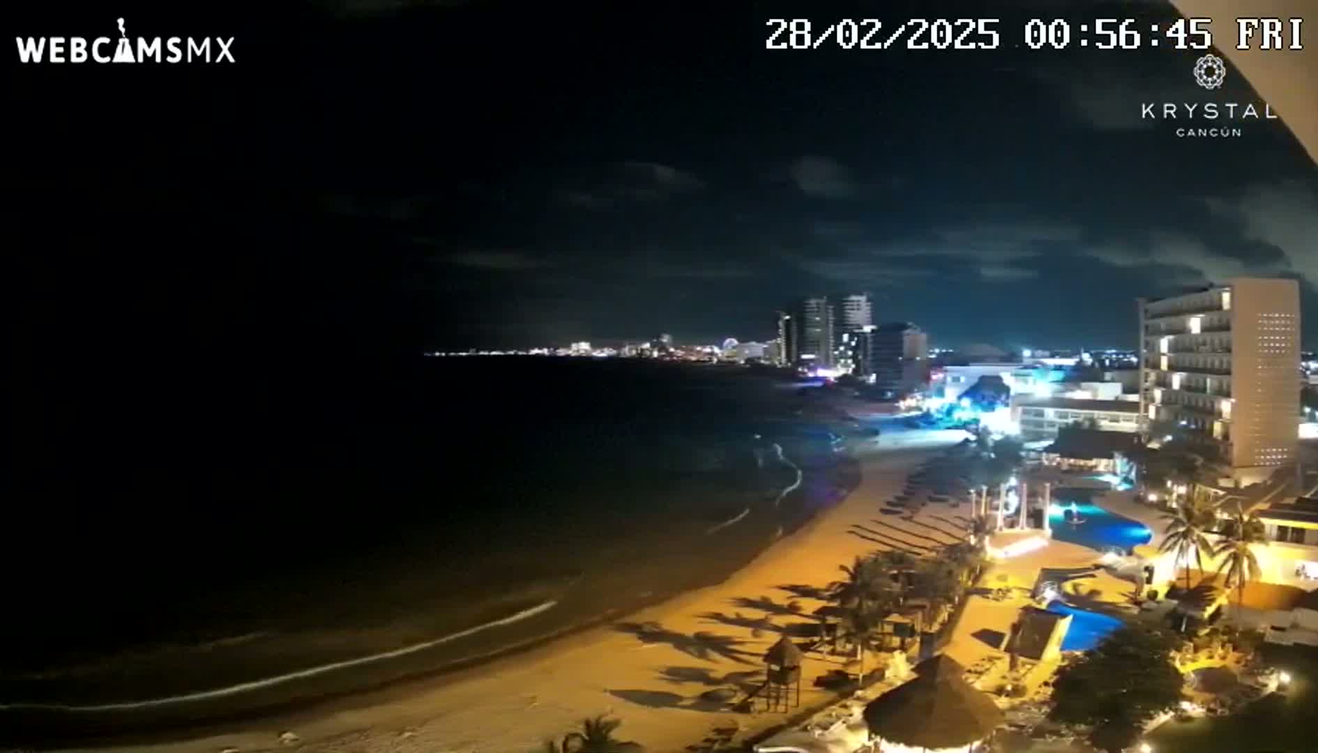 A nighttime view of a beach with calm waves, illuminated buildings, and palm trees under a partly cloudy sky.
