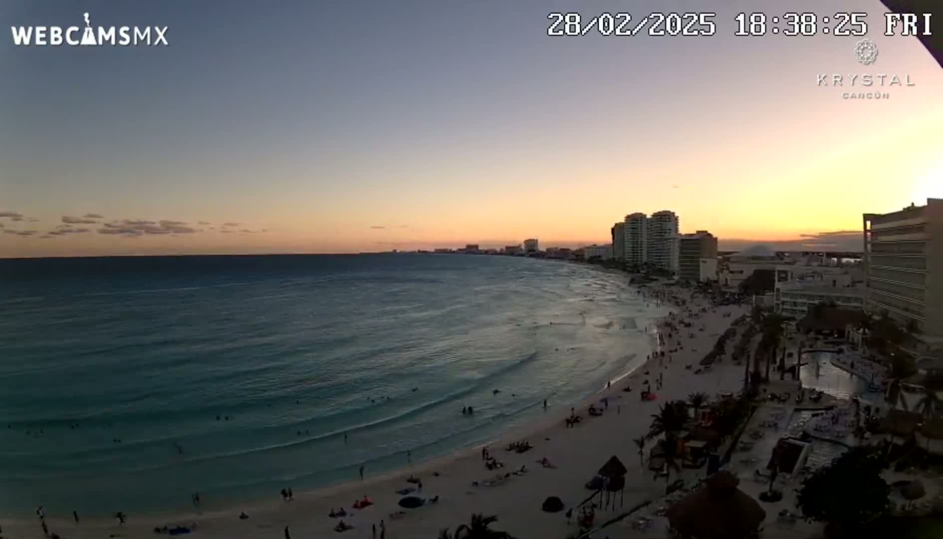 A beach with people on it and ocean waves at sunset, with hotel buildings in the background.