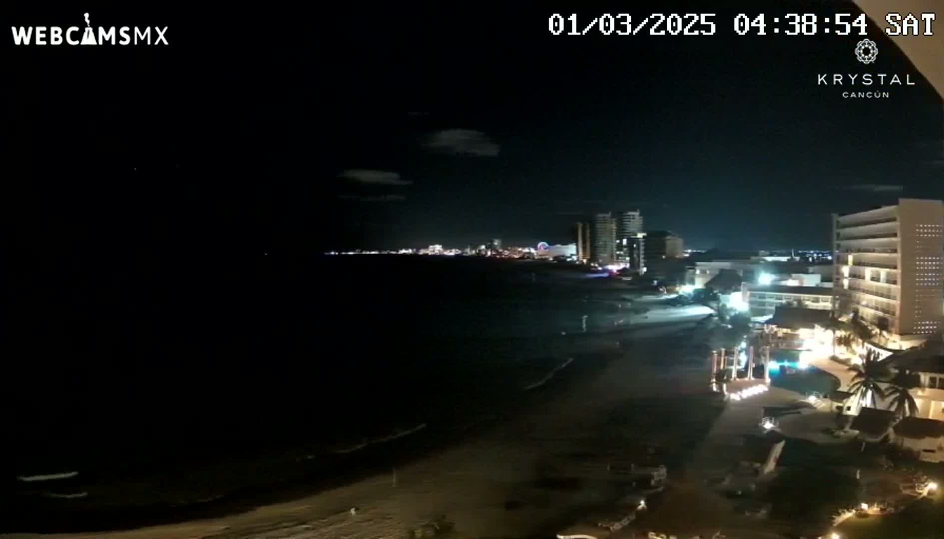 A nighttime beach scene shows a dark ocean with gentle waves lapping the shore, illuminated by the lights of a city skyline and beachfront hotels.