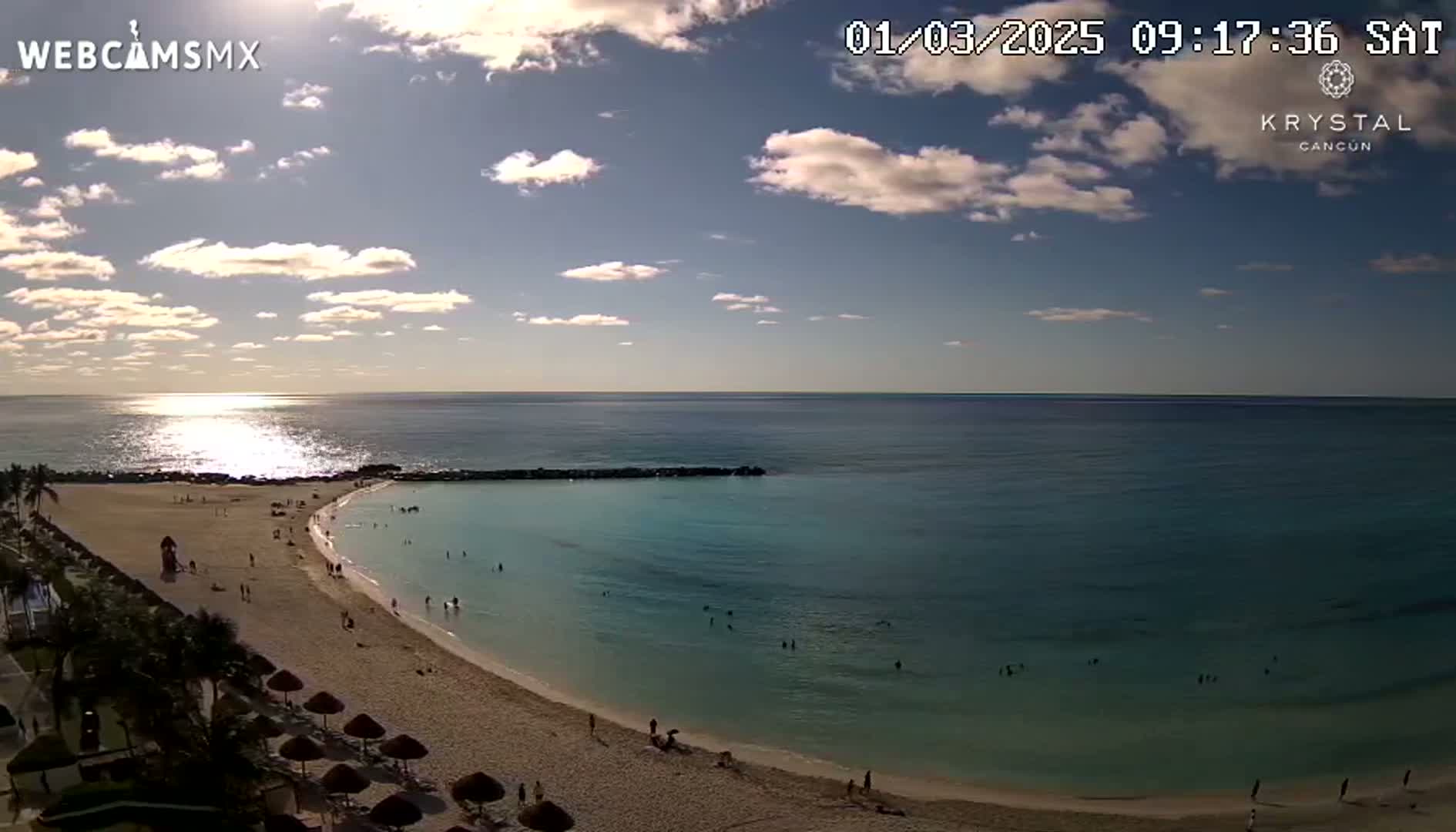 A partly sunny day shows a tranquil beach scene with calm, clear turquoise water, a sandy shore, and a few people enjoying the beach.