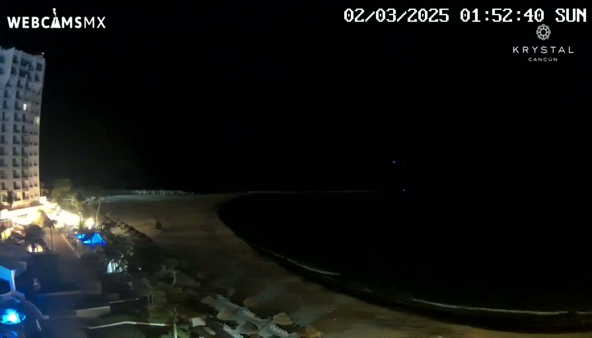 At night, a beach with a dark ocean and a tall, brightly lit building is visible.