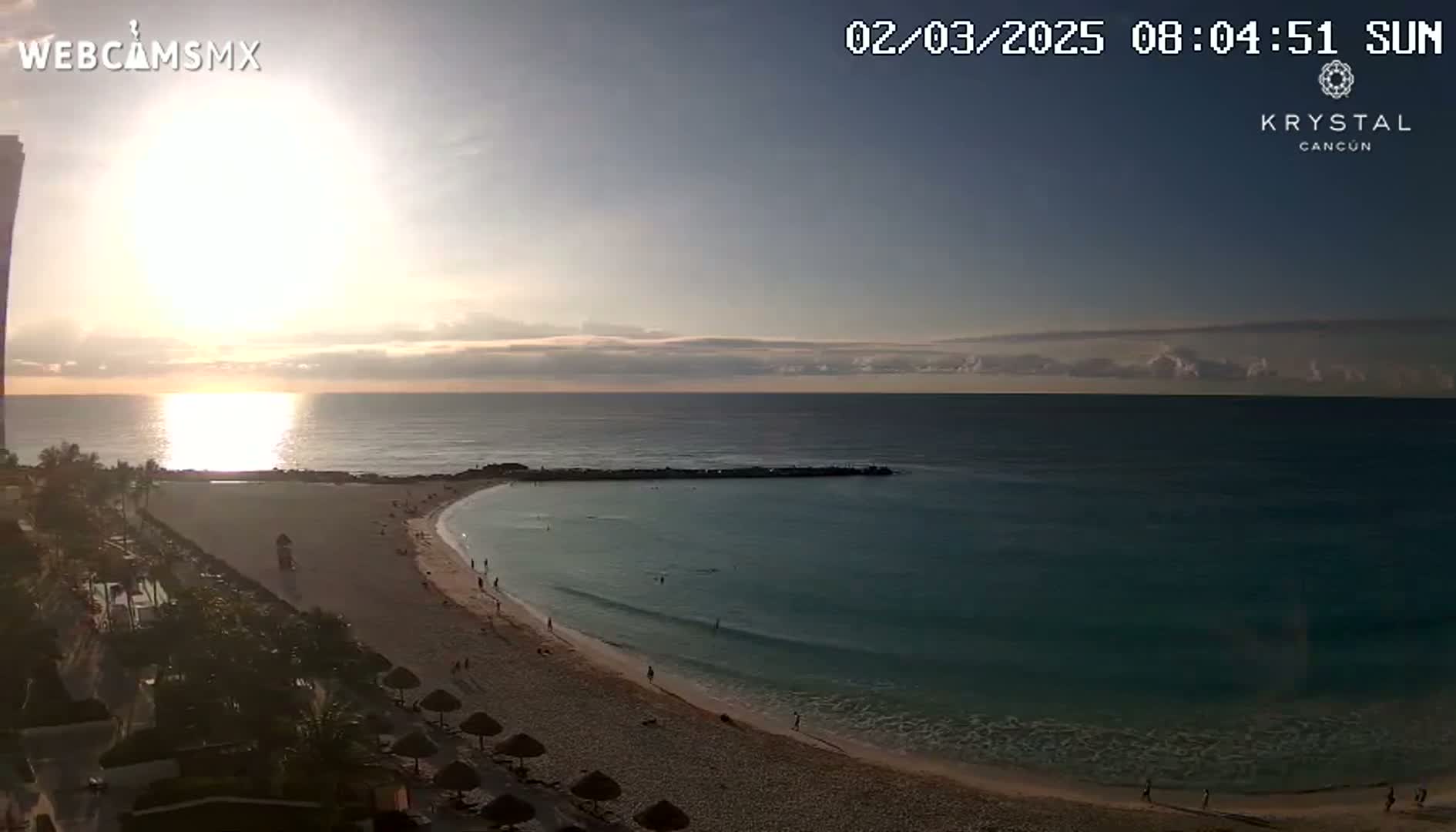A sunrise over a calm ocean illuminates a sandy beach with a few people and some beach umbrellas, near a tall building.