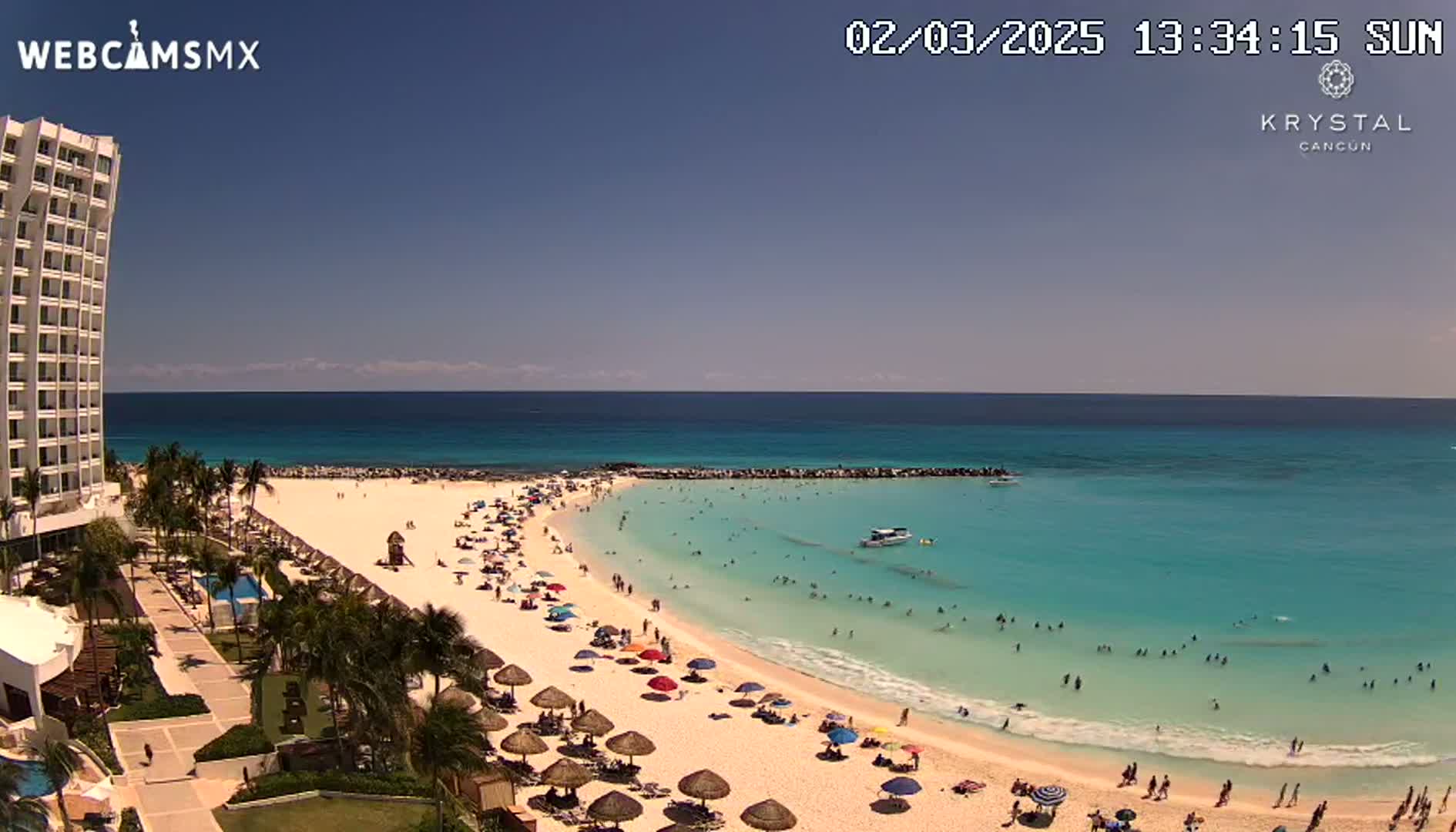 A high-angle, sunny view shows a beach with many people swimming and relaxing under umbrellas, adjacent to a tall, white hotel, and a calm, turquoise ocean.