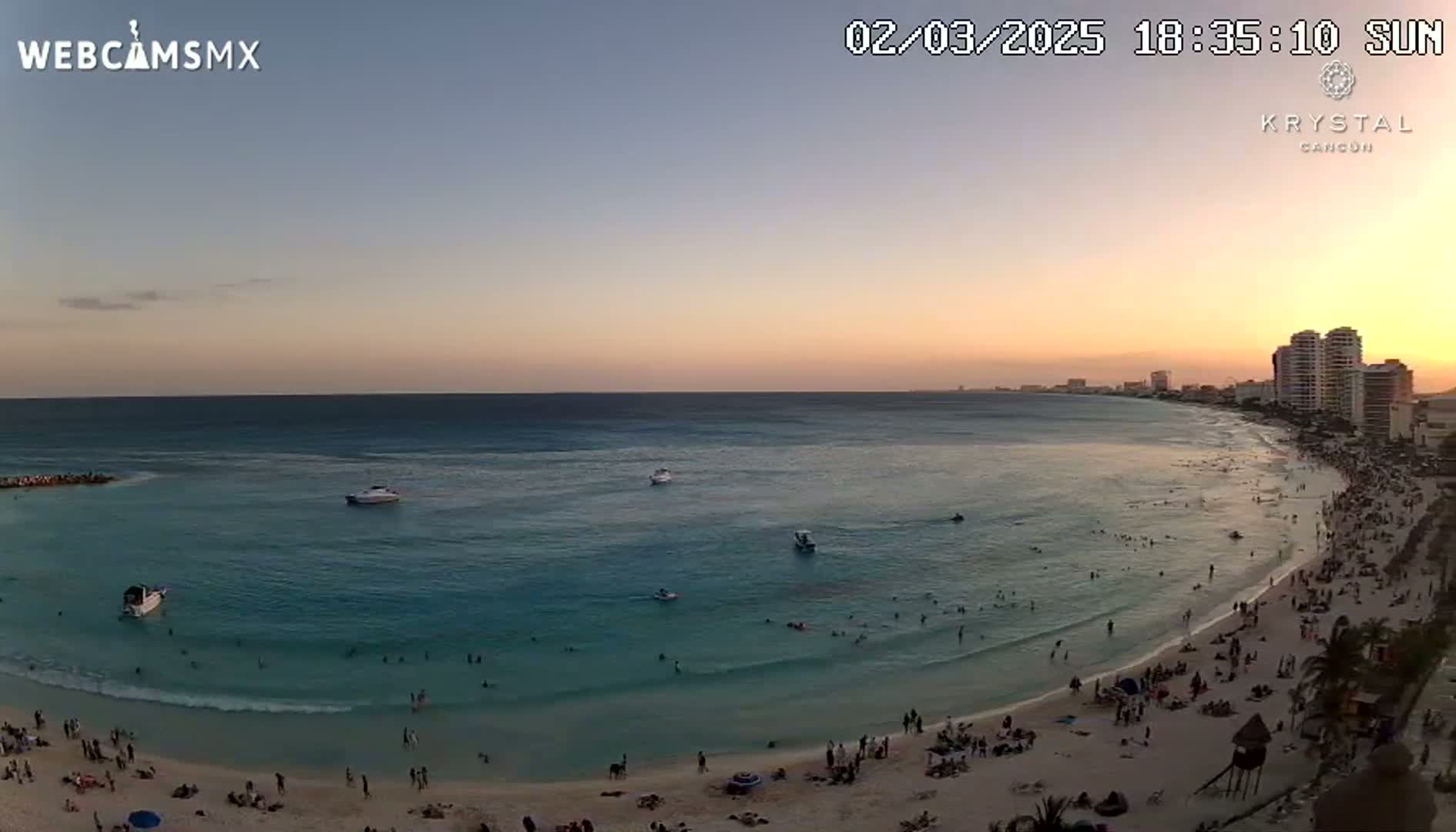 A beach at sunset with calm, clear turquoise water, many people swimming and relaxing on the sand, and several boats offshore.