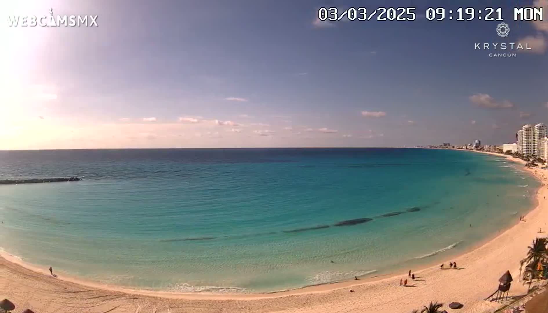 A mostly sunny day shows a long stretch of light-sand beach curving around a calm, turquoise ocean with a distant cityscape.