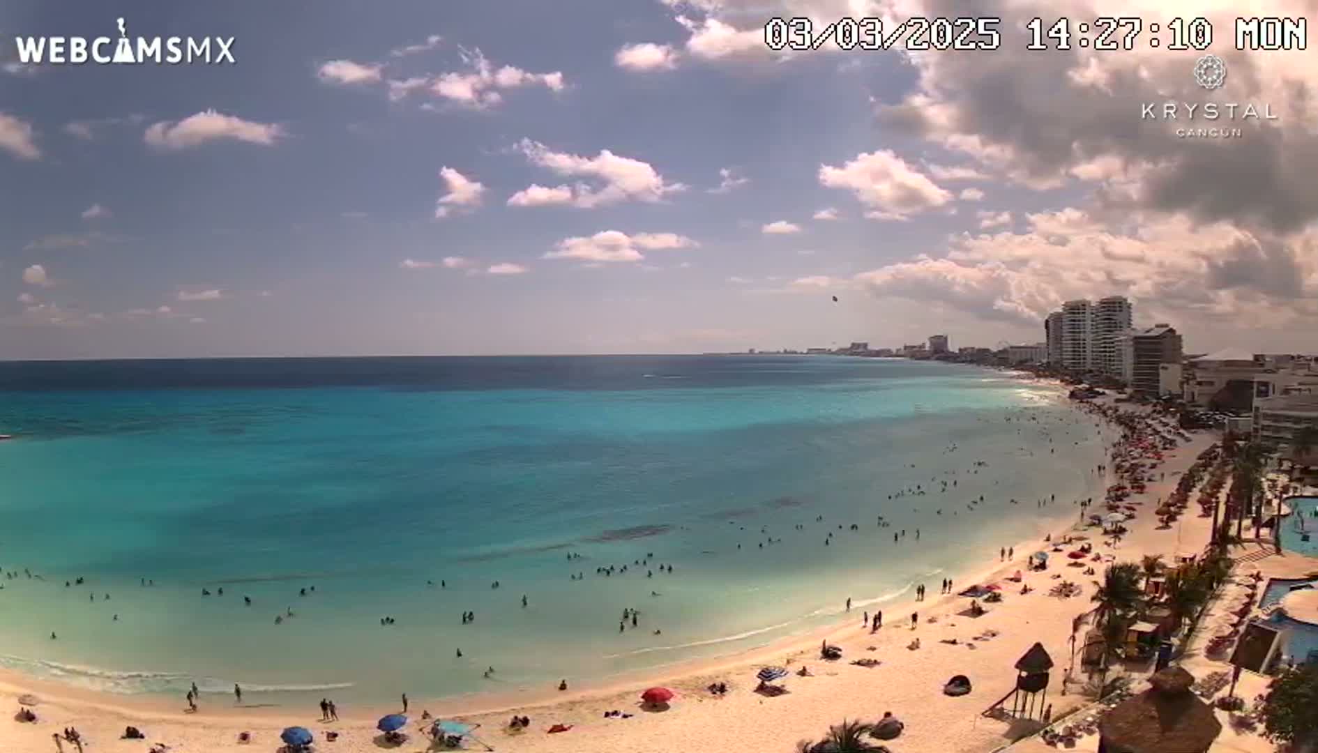 A partly cloudy day shows a beach with many people swimming and sunbathing, along a shoreline with hotels and buildings in the background.