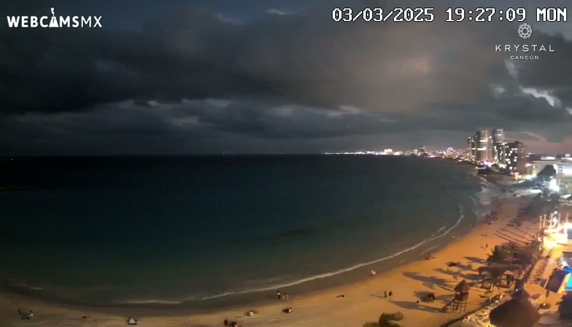 A nighttime beach scene shows a dark, cloudy sky over a calm ocean with a sandy shore, sparsely populated with people, and buildings visible in the distance.
