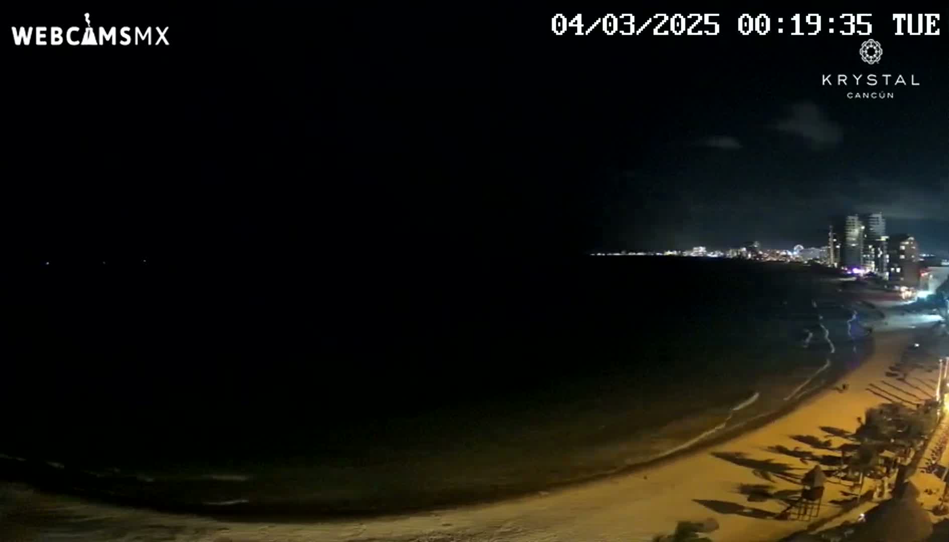A nighttime beach scene shows a dark ocean with gentle waves lapping a sandy shore, lit by artificial lights from buildings in the distance.