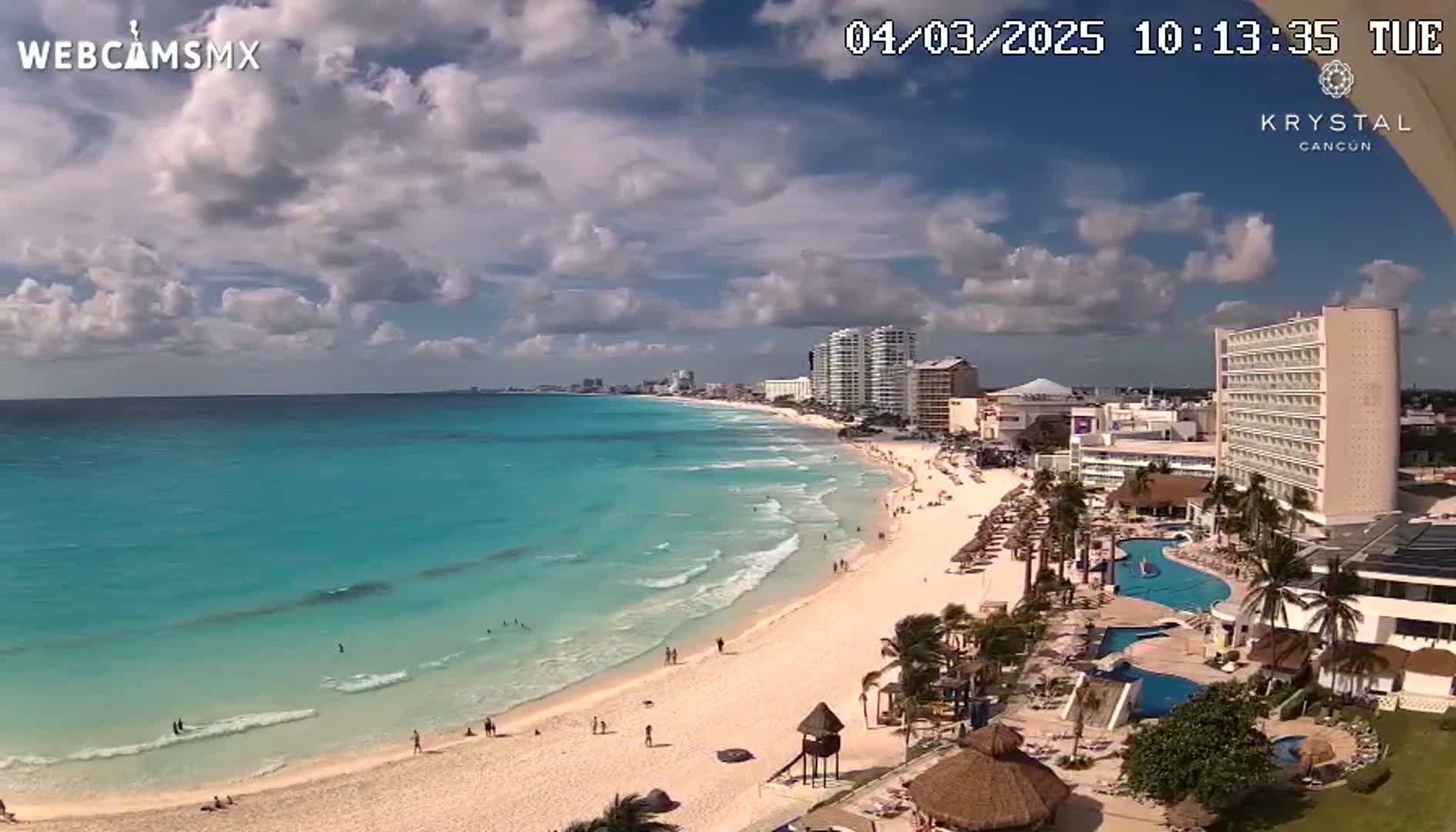 A partly cloudy day shows a long stretch of white sand beach and turquoise water, lined with hotels and resorts.