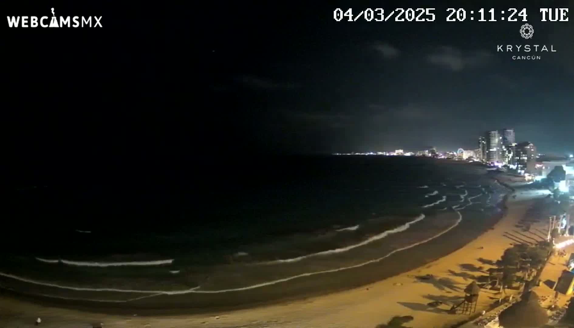 A nighttime beach scene shows gently rolling waves lapping a sandy shore, illuminated by city lights and a mostly clear sky.