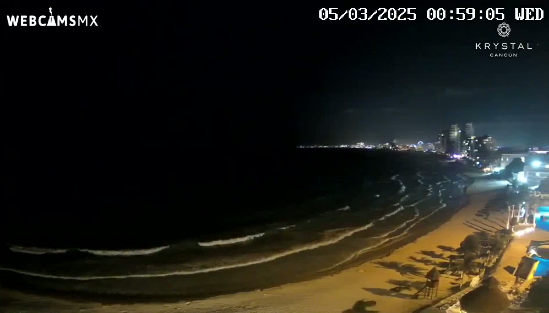 A nighttime beach scene shows gently rolling waves lapping a sandy shore, with city lights visible in the distance under a dark sky.