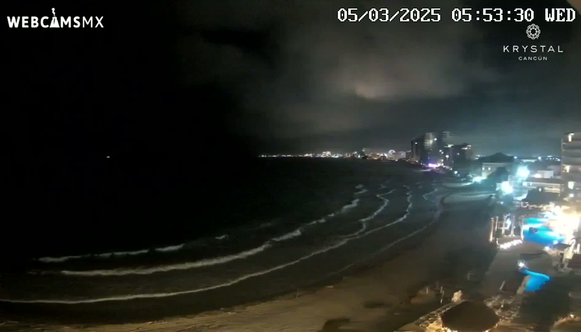 A nighttime beach scene shows gently rolling waves on a dark ocean under a mostly cloudy sky, with city lights visible in the distance and illuminated buildings along the shore.