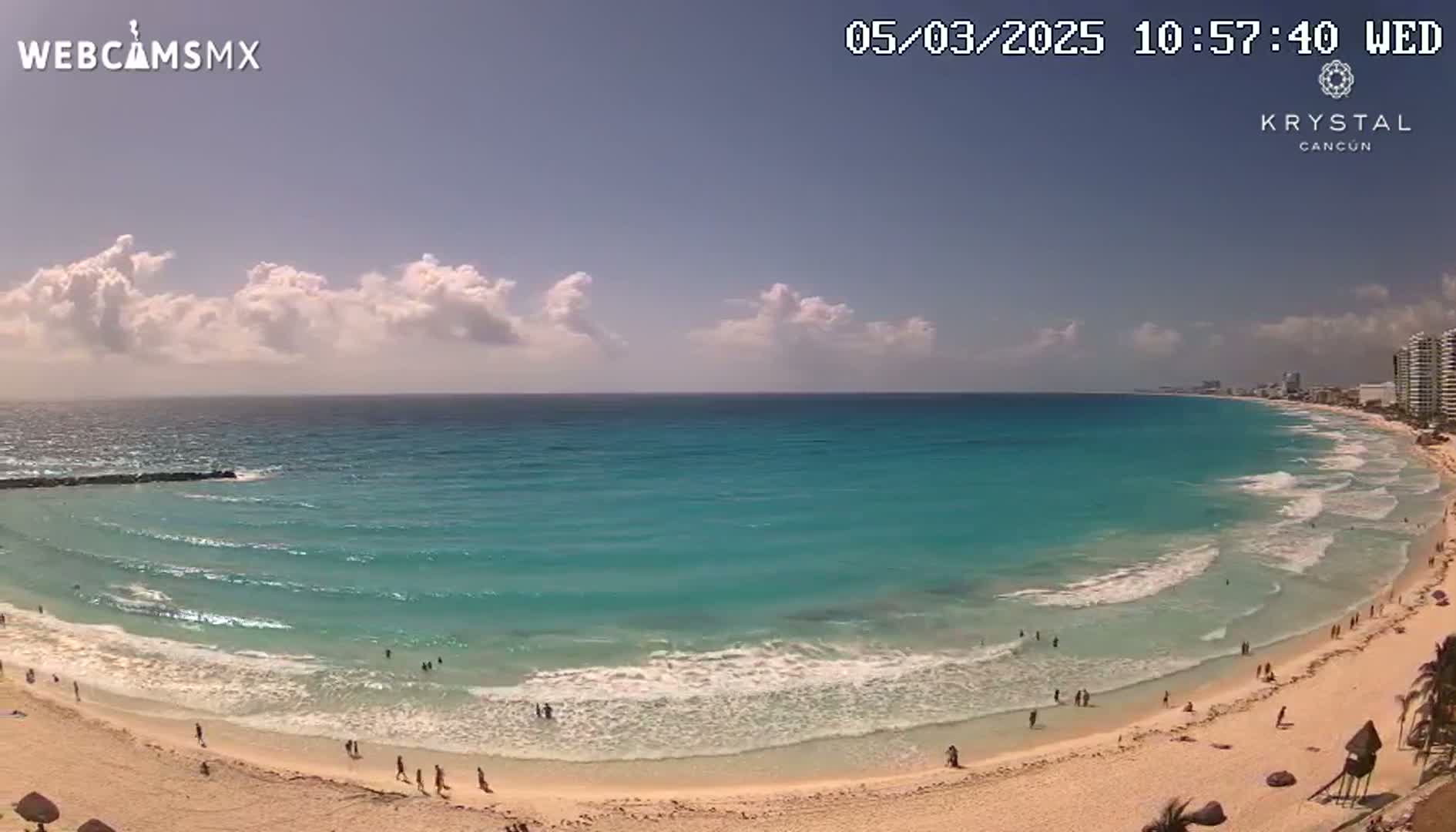 A partly cloudy day shows a long, sandy beach with turquoise water and gentle waves, populated by a few people.