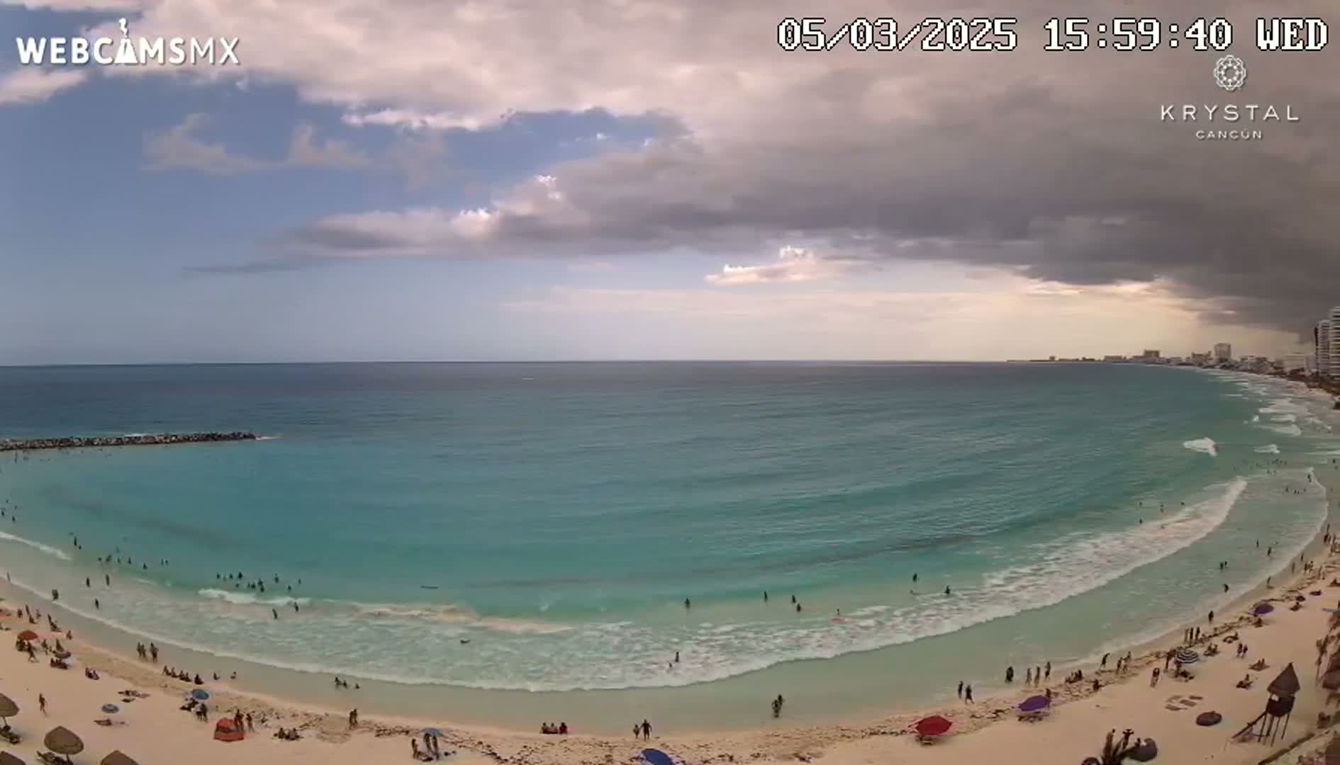 A partly cloudy sky hangs over a beach with turquoise water, where people swim and relax on the sand.