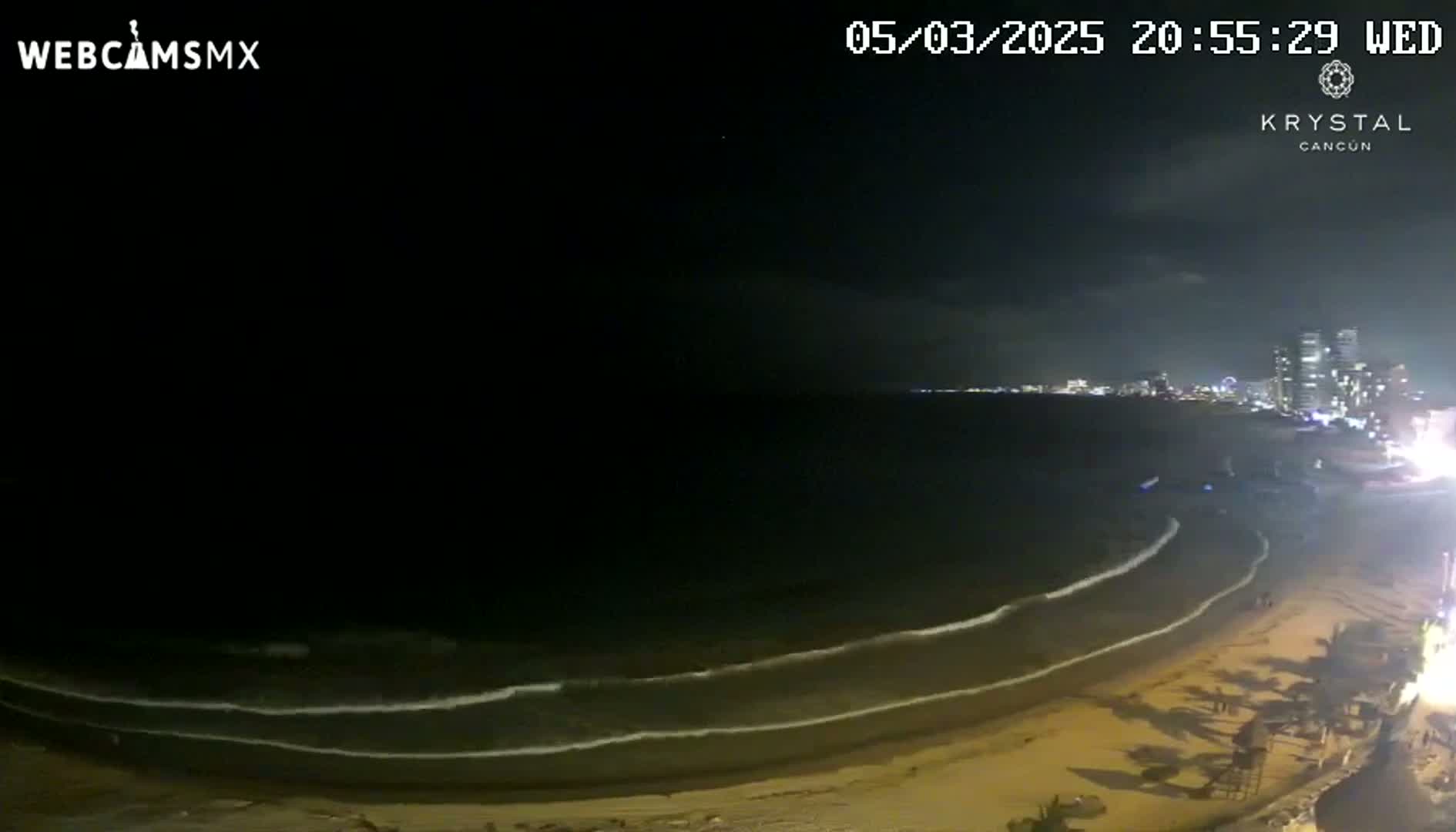 A nighttime beach scene shows gently lapping waves on a sandy shore with a city skyline visible in the distance under a dark, clear sky.