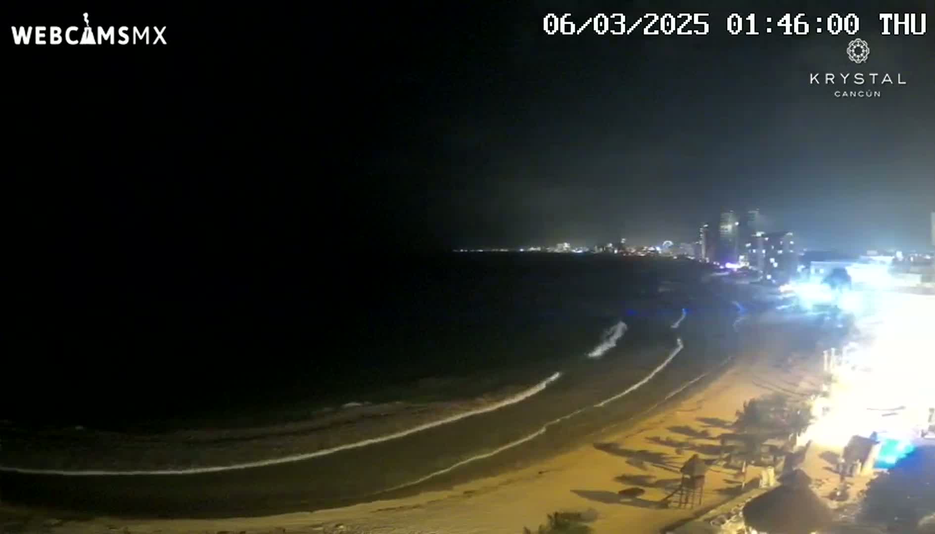 A nighttime beach scene shows gently lapping waves on a sandy shore, with a city skyline illuminated in the distance under a dark sky.