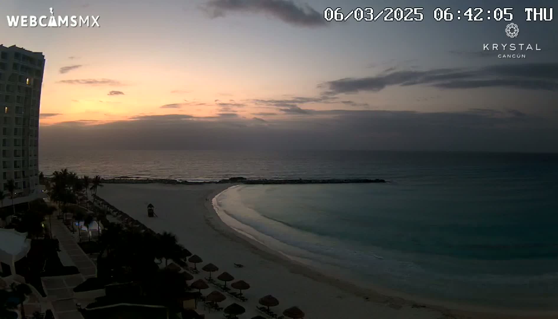 A beach with calm ocean waters is seen at sunrise, next to a tall hotel.