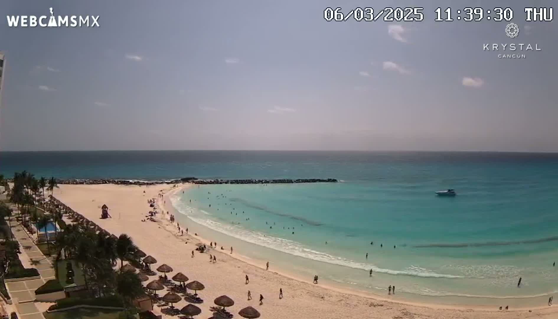 A partly sunny day shows a sandy beach with turquoise water, palm trees, and beach umbrellas, with a few people swimming and relaxing.
