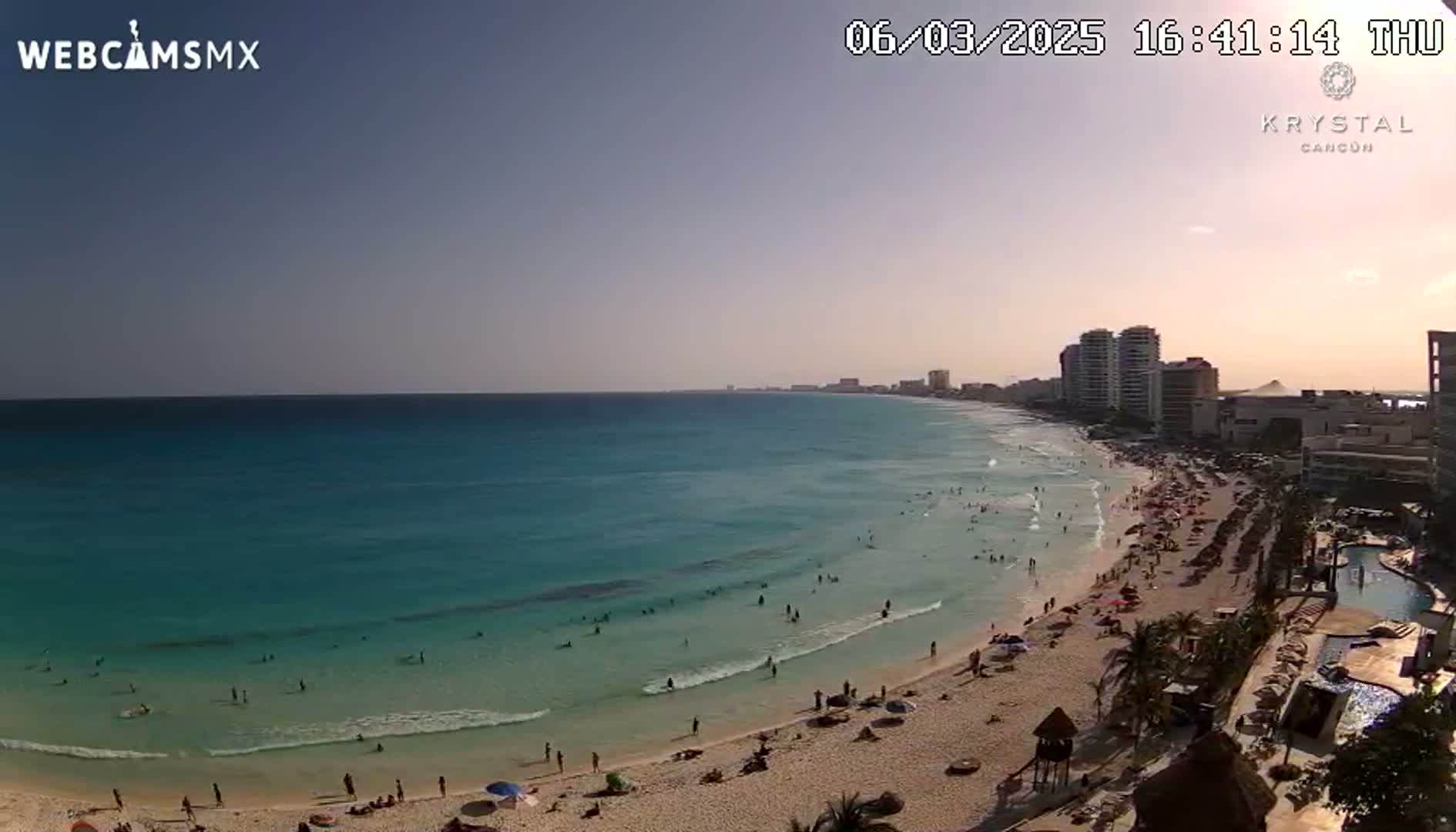 A beach scene on a partly sunny day shows turquoise water with people swimming, a sandy shoreline with sunbathers, and high-rise hotels in the background.