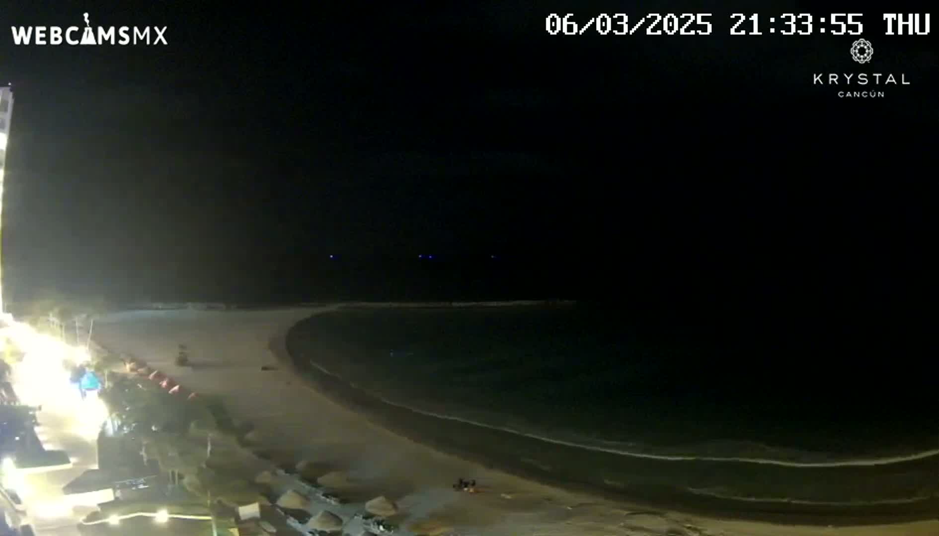 A nighttime view of a dark beach with calm waves and dimly lit buildings along the shore.